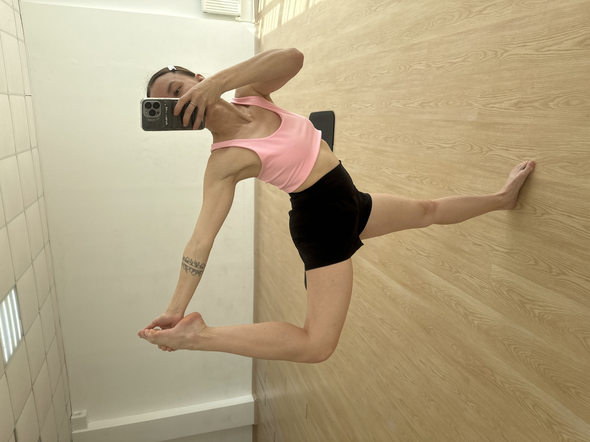 A person performs a challenging yoga or flexibility pose indoors, captured in a selfie taken with a smartphone. The individual, dressed in a pink sports bra and black shorts, stands on their right leg while extending their left leg backward and upward, bending at the knee, and reaching back with their left hand to grasp the left foot. Their right hand holds an iPhone, partially obscuring the face, as they look towards the camera. A tattoo with a circular pattern is visible on the left forearm. The setting features a light wooden floor and white walls; one wall exhibits a grid of square white tiles, while another is plain white. Natural light illuminates the room, suggested by shadows on the floor. No clearly legible text is visible within the image itself. This image was taken in Votkinsk, Russia.