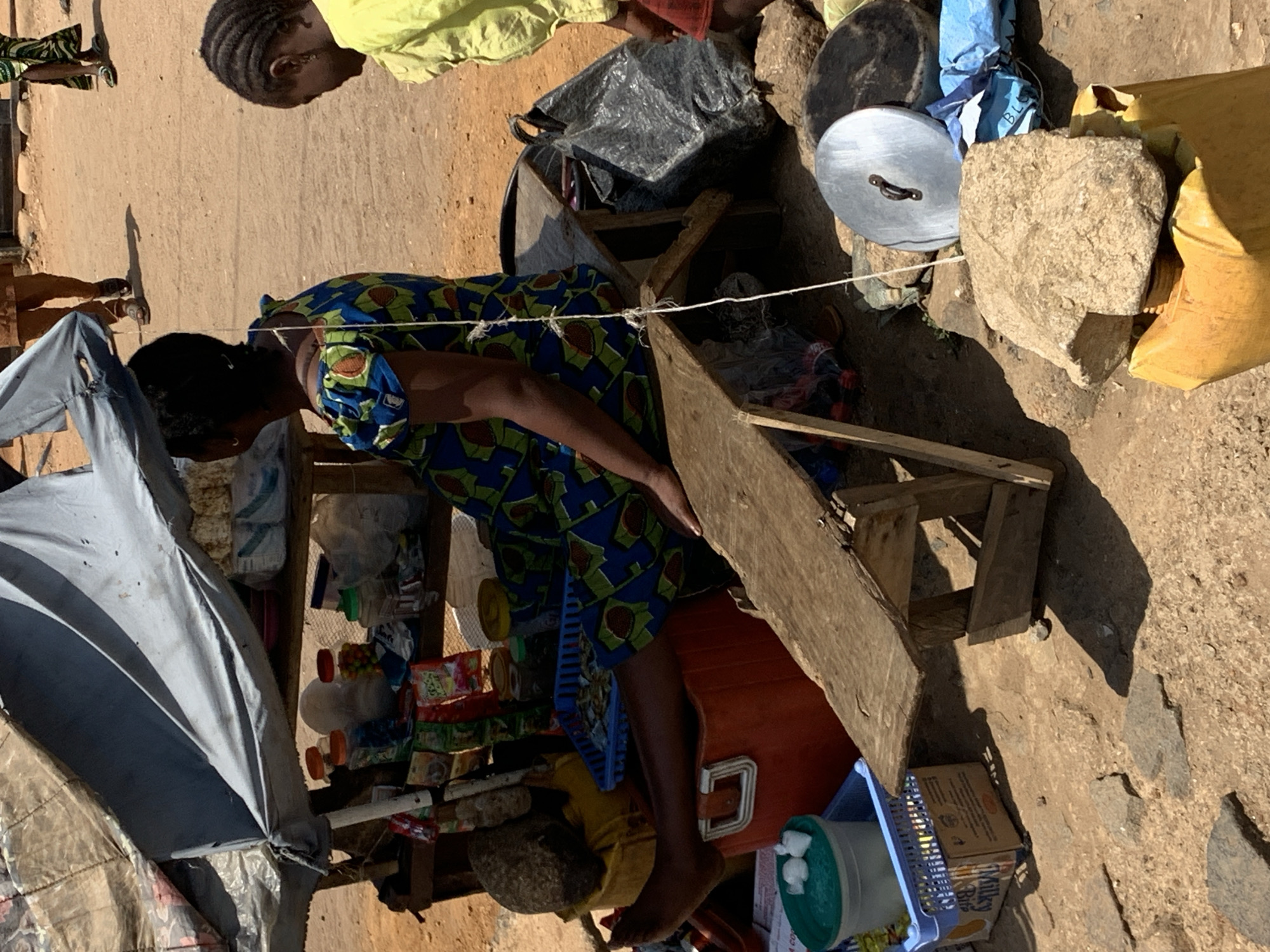 A woman wearing a patterned dress is operating a roadside stall in Rafin Par, Nigeria. The stall is constructed of wood, an orange cooler, and other items. Assorted goods for sale are displayed. A blue and white umbrella provides shade. Two additional people are present, one standing and one near the top of the frame. The ground is dirt and stone.