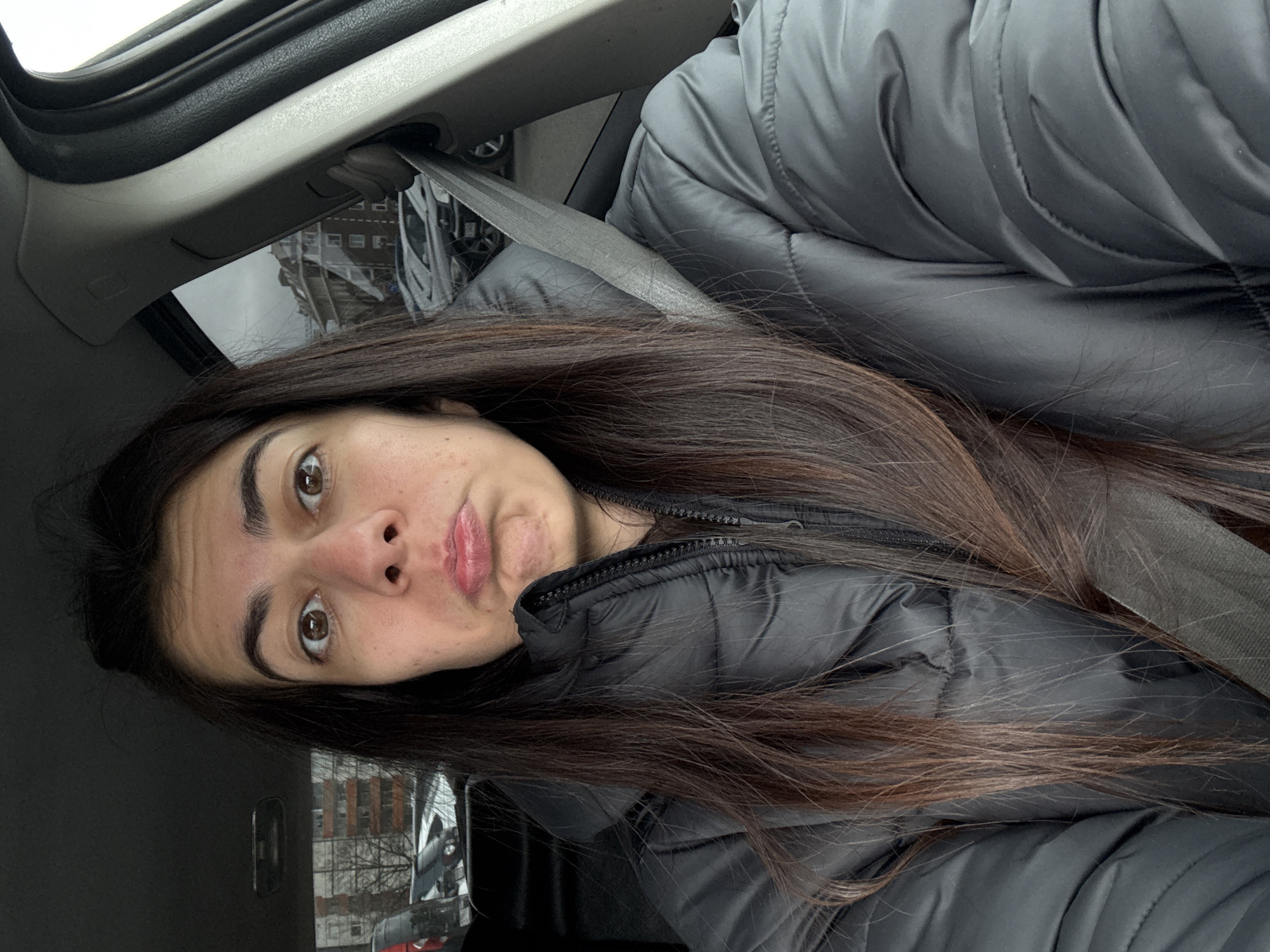 A woman is seated in a vehicle, wearing a padded, grey coat and a seatbelt. She is looking at the camera with a slight pout. The background outside the car window includes buildings and trees, possibly located in Sofia, Bulgaria. The scene appears to be taken during the daytime.