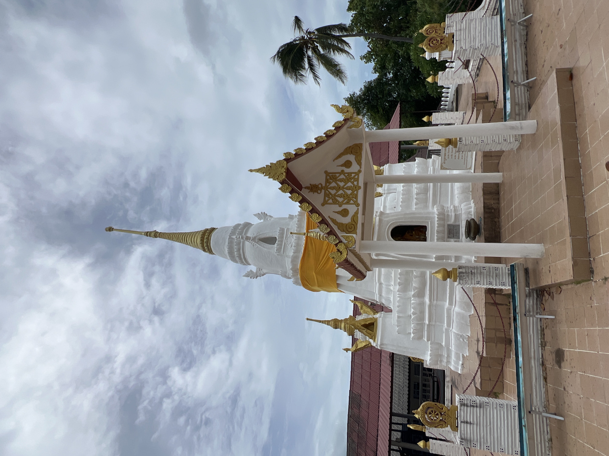A white, multi-tiered Buddhist pagoda with a gold spire stands prominently in the foreground of an overcast day in Ko Samui District, Thailand. A bright yellow sash is wrapped around the upper section of the pagoda. In front of the pagoda, a smaller white structure with ornate gold trim and roof details is visible, supported by white pillars. The ground is paved with tan tiles, and a low chain barrier surrounds the pagoda area. To the left and right of the pagoda, white pillars topped with gold finials and decorative gold emblems are present. In the background, a red-roofed building is partially visible, and a lush green forest with a tall palm tree swaying in the wind completes the scene. The sky is filled with a mix of white and gray clouds.