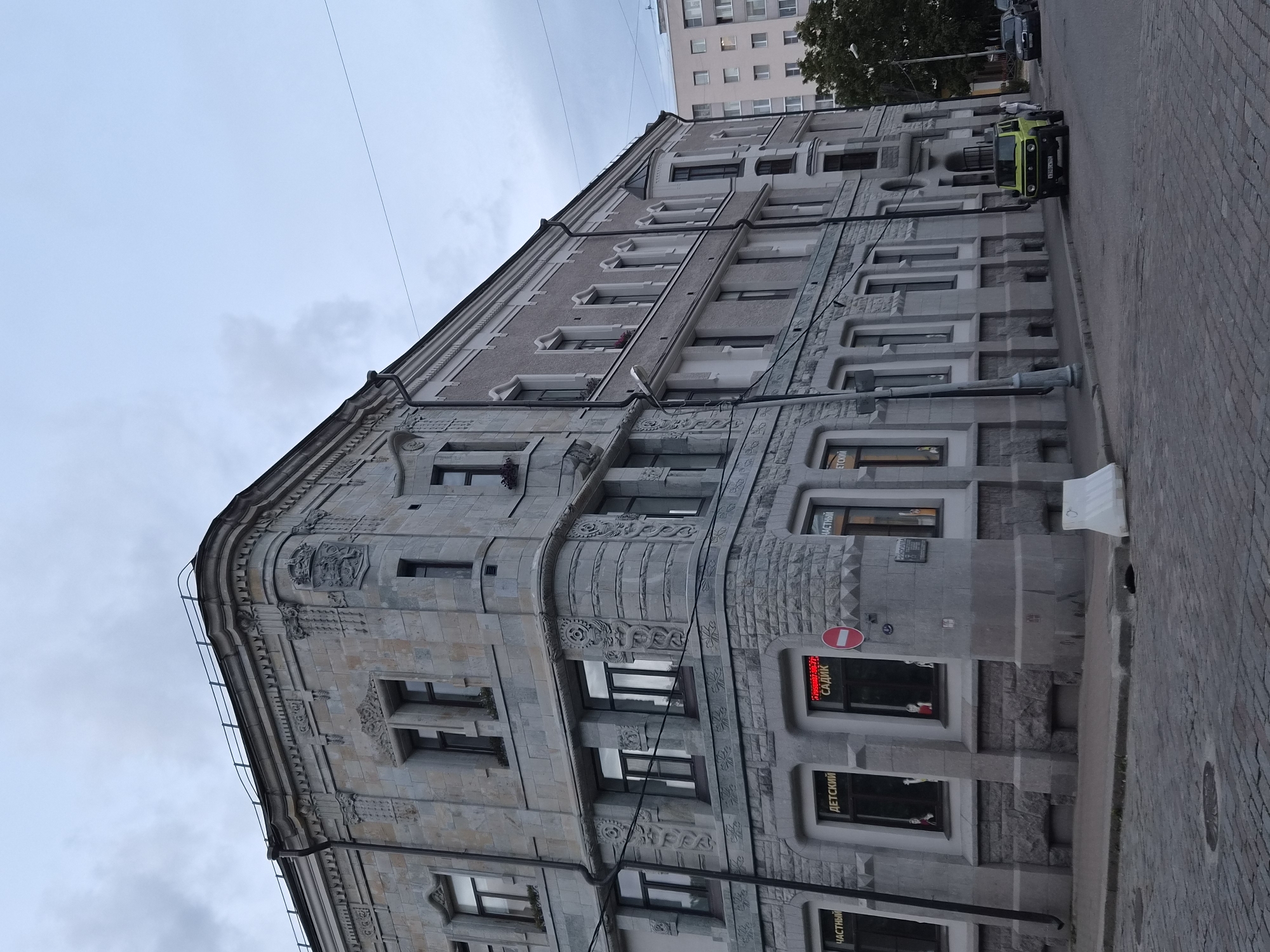 A multi-story building with ornate stone facade and a lighter upper section is depicted from a low-angle, street-level perspective. The building has numerous windows, some of which feature decorative stonework. On the ground floor, businesses are visible, including one with a red neon sign reading "САДИК" (Sadik) and another with "ДЕТСКИЙ" (Detskiy), indicating children's services. A red circular sign with a white bar signifies a prohibition. A light green SUV is parked on the cobblestone street in front of the building, with a person walking behind it. A white traffic barrier is present on the sidewalk. In the background, a modern apartment building and trees are visible under a cloudy sky. The scene is set in the city of Vyborg, Russia.
