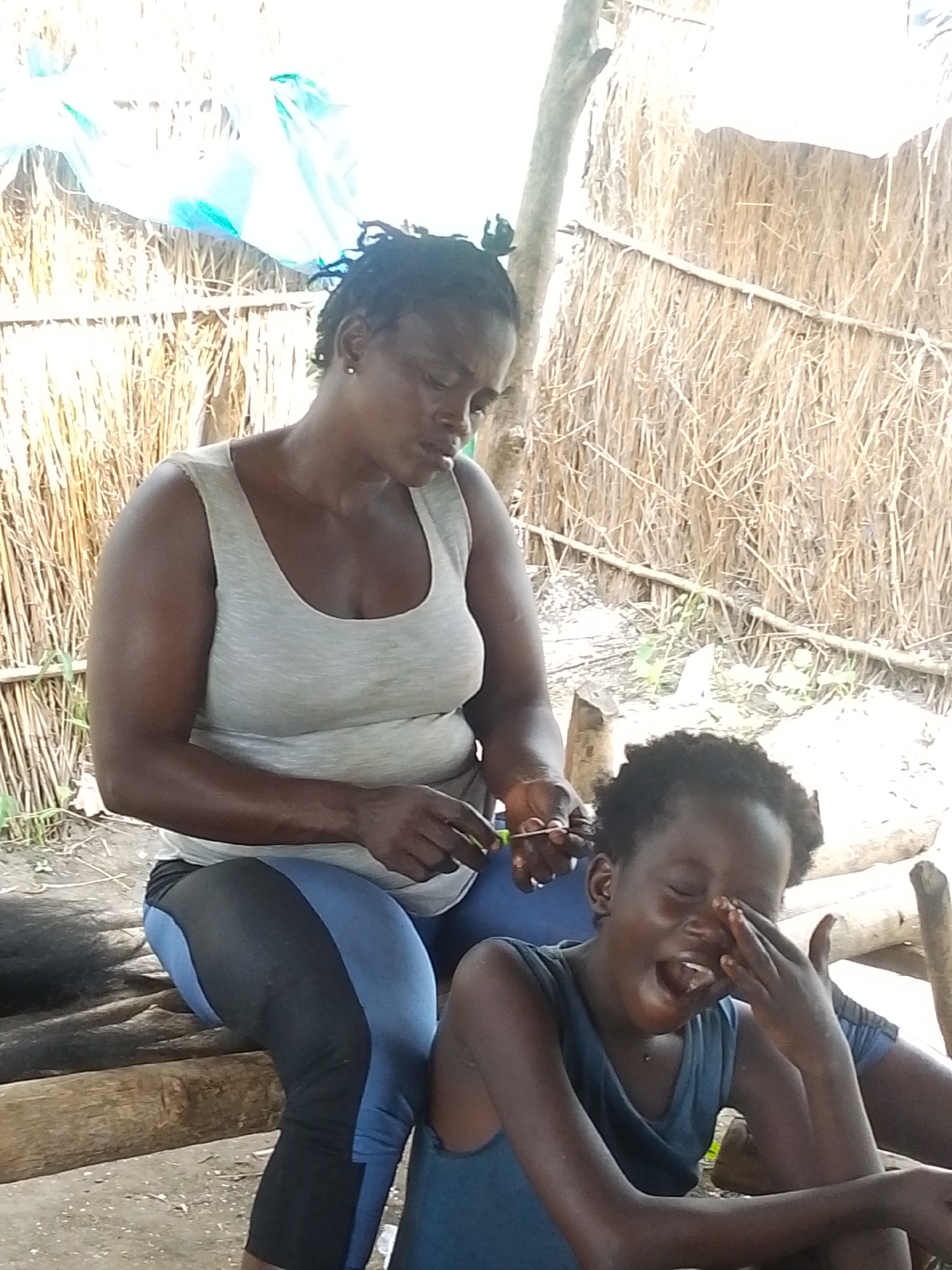 A Black woman with dark skin and braided hair is braiding the hair of a Black child. They are sitting on a rough wooden bench outdoors in what appears to be a rural or semi-rural setting, possibly in Otulu, Nigeria. The woman wears a light grey tank top and blue and black leggings. She is using a comb and her hands to style the child's hair. The child, with dark skin and short, curly hair, is wearing a blue tank top and is leaning back, with their eyes closed and mouth open, appearing to be either crying or yawning. The background consists of a wall made of dried straw or reeds, with some dry branches and what looks like a blue tarp draped overhead. The ground is a mixture of dirt and gravel. The lighting suggests it is daytime.