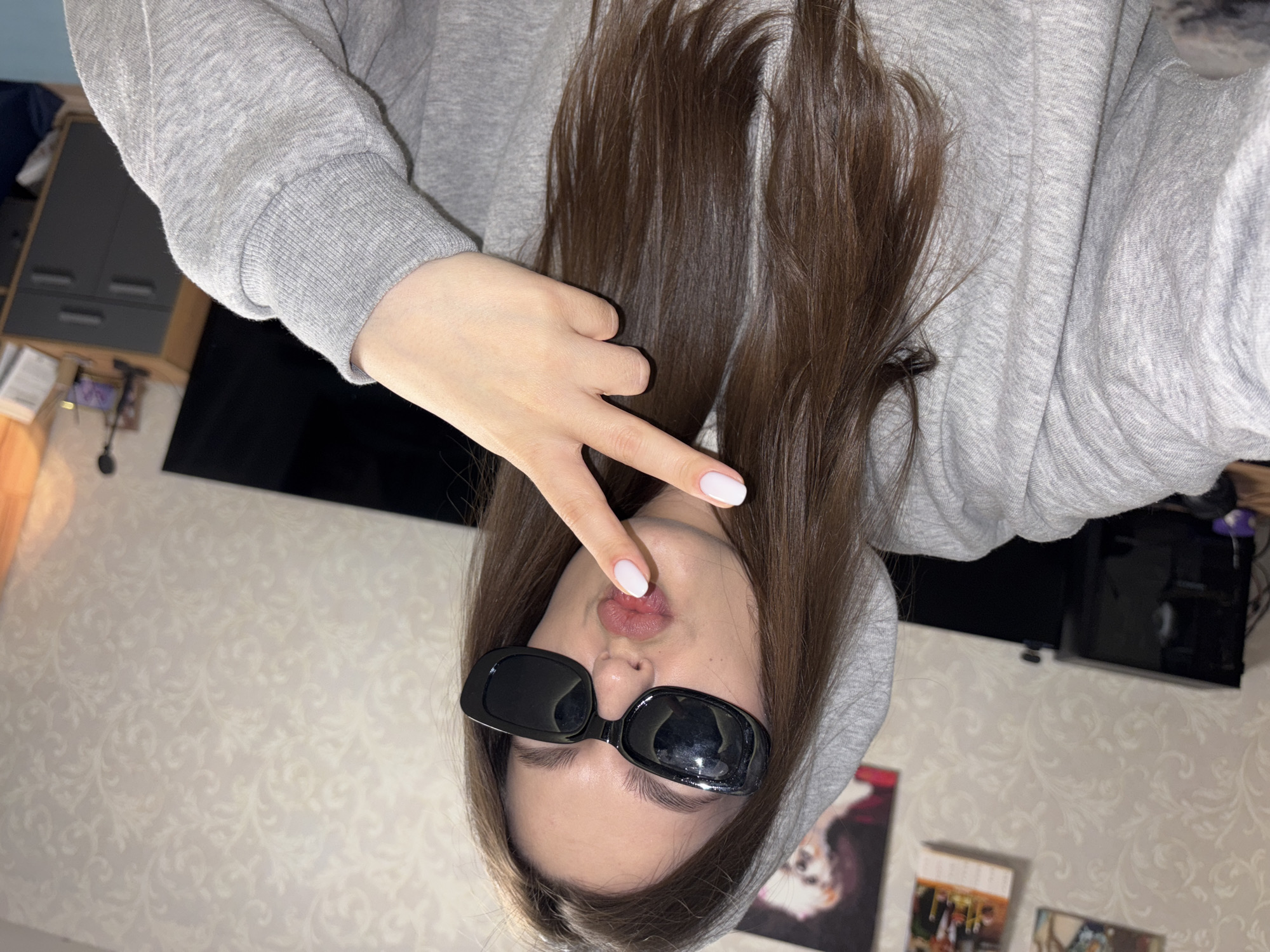 This media file features a person, likely female, captured in a close-up, selfie-style photograph. The image is oriented ninety degrees counter-clockwise, showing the person appearing upside down. The subject is wearing a light grey hooded sweatshirt (hoodie) with visible texture on the fabric. They have long, straight, dark brown hair that frames their face and extends around their shoulders. Black, rectangular sunglasses cover their eyes. Their lips are pursed into a "duck face" or "kissy face" expression. One hand is raised near their face, making a "V" or peace sign, with fingernails painted a clean white.
The setting is indoors, characteristic of a residential space in Chaikovsky, Russia. The background reveals light-colored wallpaper adorned with a subtle, ornate damask-like pattern. Visible elements of the room include a dark grey cabinet or piece of furniture to the left, and a dark, rectangular electronic device (possibly a television or monitor) mounted or placed on a surface in the distance. Other indistinct objects and framed pictures or posters are also partially visible on the walls and surfaces, suggesting a lived-in environment. The lighting appears to be artificial, typical of indoor illumination. The overall activity depicted is a casual pose for a photograph, conveying a playful or relaxed mood. No discernible text is visible within the image.