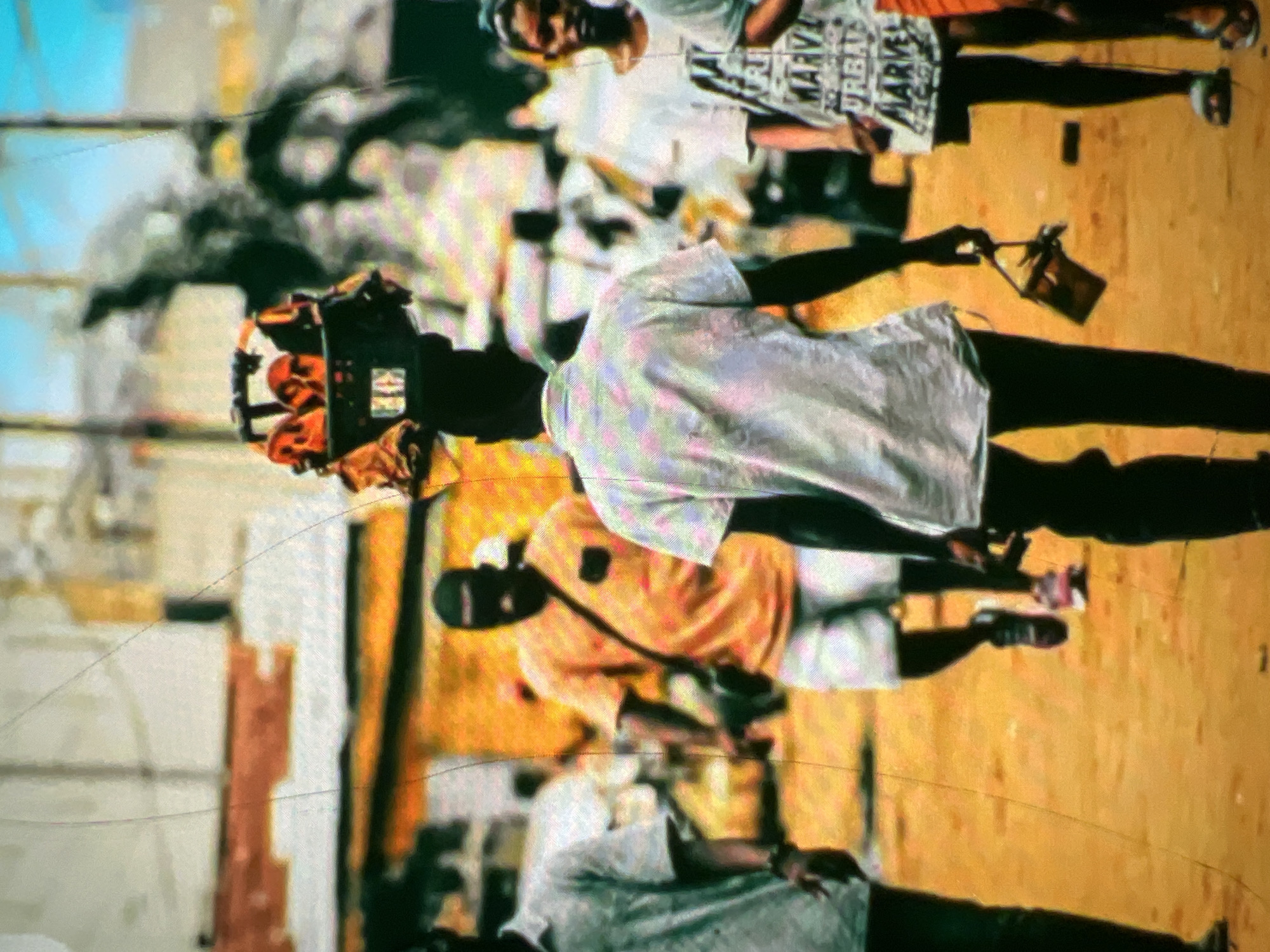 In Lagos, Nigeria, a street vendor balances a crate filled with fruit on their head, walking forward through a dusty, sun-drenched street. The vendor is seen from behind, wearing a light-colored, patterned shirt and dark pants. A small, dark bag hangs from their right hand. Other pedestrians are visible in the background, including a man in an orange shirt and white shorts walking away from the camera, and a woman in a white t-shirt with text and a girl in a light-colored shirt to her right. The environment appears to be a bustling urban area with buildings and a road surface composed of reddish-brown dirt.