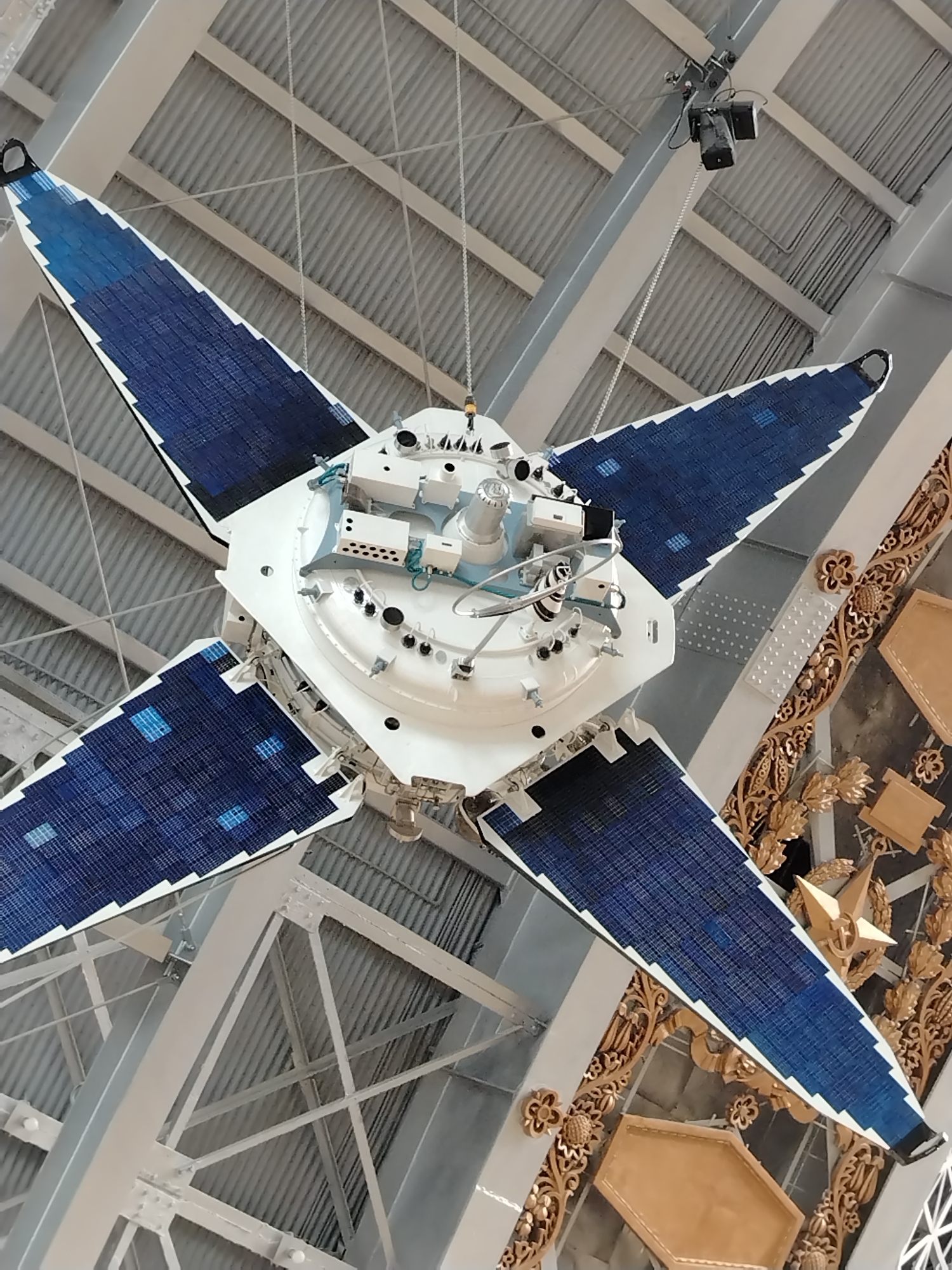 The image shows a detailed view of a satellite model suspended from the ceiling of a large indoor space. The satellite has a central white body with various scientific instruments and sensors attached. It is equipped with four large, wing-like solar panels, each covered in blue solar cells arranged in a zig-zag pattern. The solar panels are angled outwards from the central body.
The background reveals a vast industrial-style ceiling with exposed metal beams and corrugated metal panels. Multiple thin cables are visible, suspending the satellite. In the upper right portion of the image, a camera is mounted on a bracket attached to a ceiling beam. The satellite is a prominent object in the foreground.
The overall impression is of a museum exhibit or a science center displaying space exploration technology. The ornate golden decorative elements on the right side of the image suggest a historical or perhaps governmental context for the display.
While the image itself does not explicitly provide location information, the presence of ornate golden decorative elements, particularly those that appear to be Soviet-era or Russian state symbols (like the star and laurel wreath elements), and the style of the architecture, could suggest a location within Moscow, Russia, possibly a museum dedicated to space or aviation. For example, the Museum of Cosmonautics in Moscow features such exhibits and decorative styles.