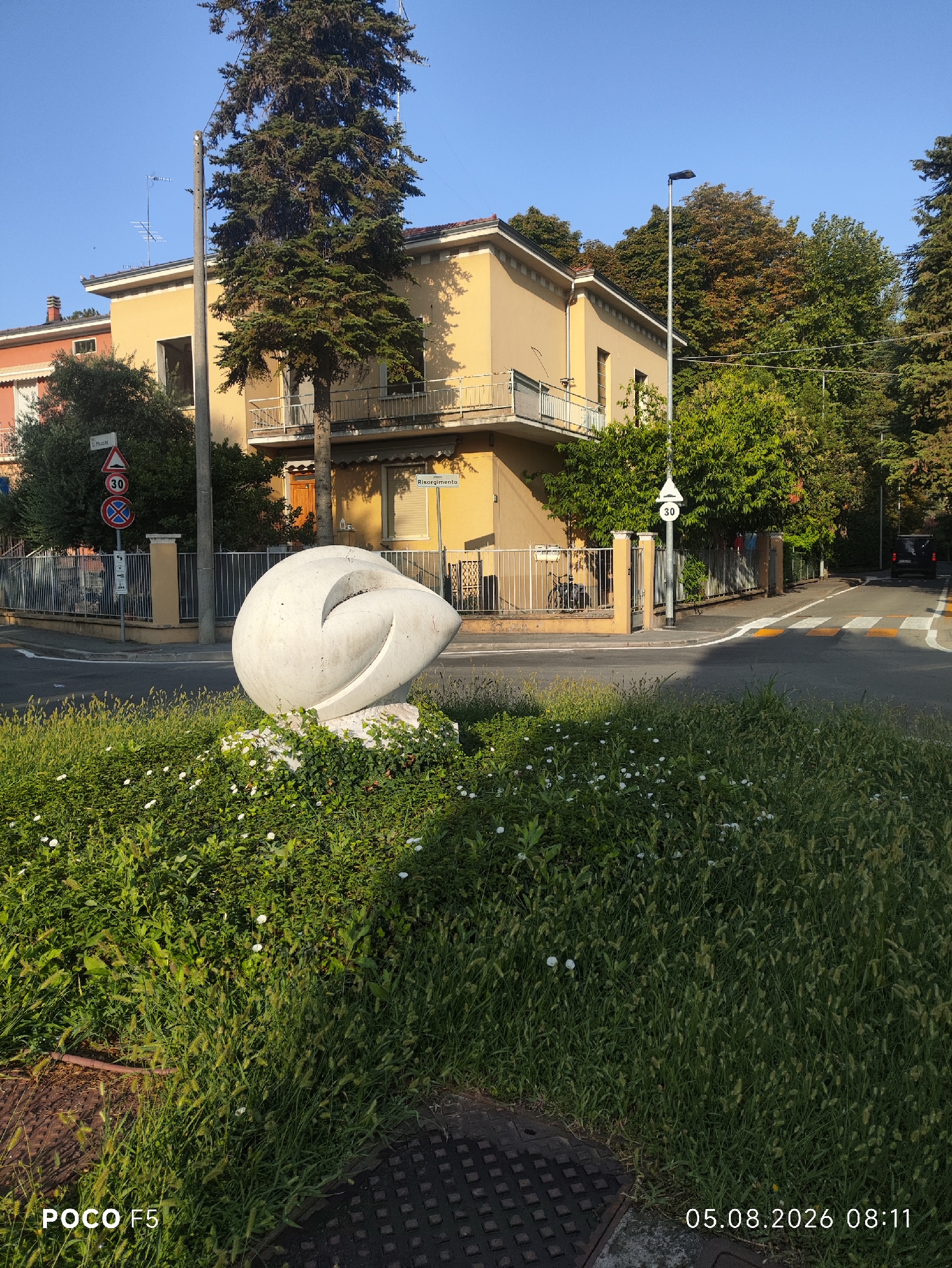 A concrete sculpture resembling a large, abstract shell is situated in a bed of green foliage with small white flowers. The sculpture is positioned in the foreground, to the left of the image. Behind it, a paved road curves. Across the road, a light-yellow building with balconies and a dark wooden door is visible. A tall evergreen tree stands next to the building. Several traffic signs are present: a "30" speed limit sign with a red circle and a white background, and another "30" sign on a triangular white background. A street lamp with a dark rectangular head stands near the building. The scene appears to be in a residential area, with a metal fence bordering the property. The sky is clear and blue. The image captures a view from street level looking slightly upwards towards the building and trees. The date and time stamp "05.08.2026 08:11" is visible in the bottom right corner, along with "POCO F5" in the bottom left corner. The location is identified as Collecchio, Italy.