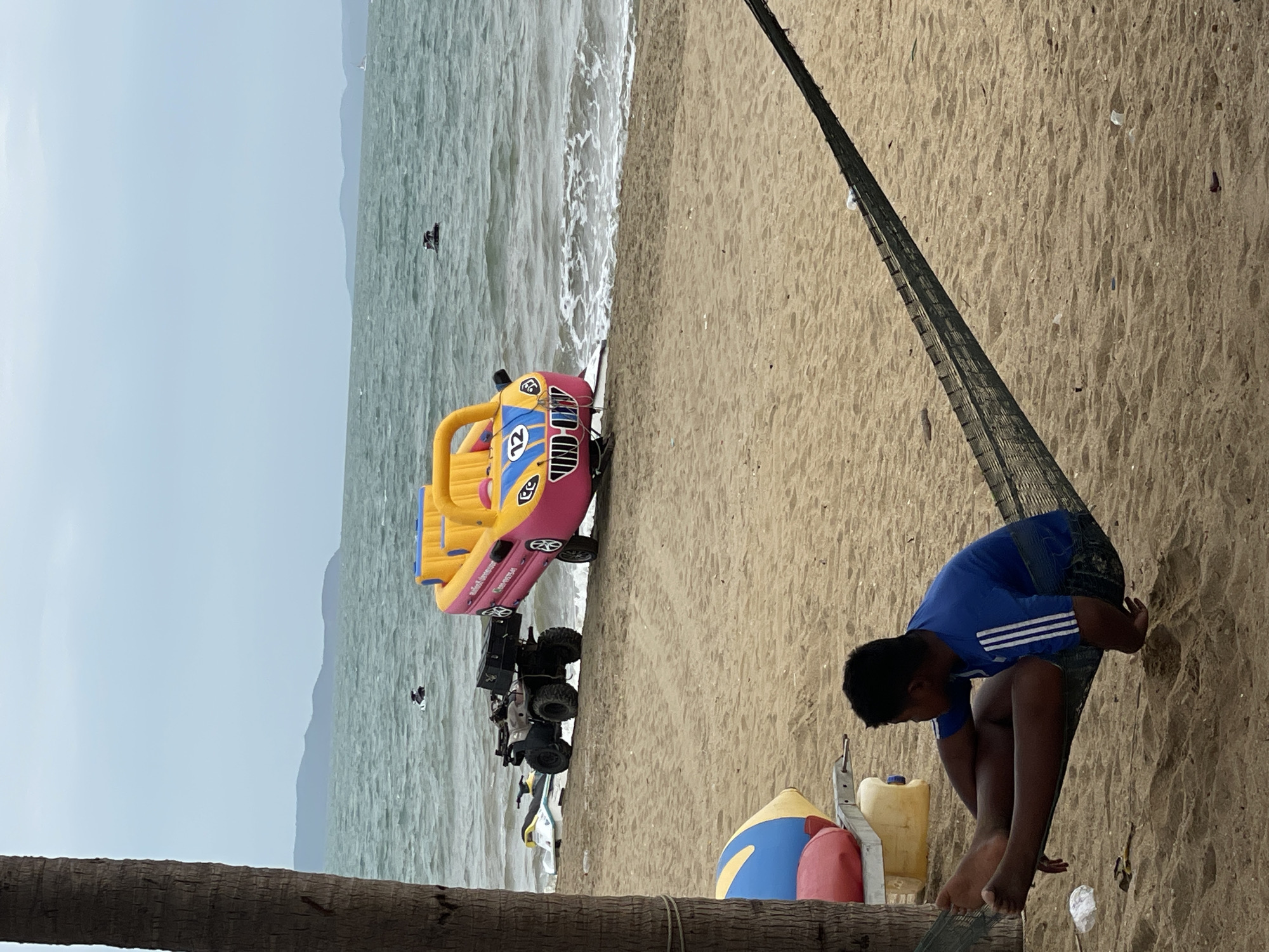A boy is relaxing in a hammock on a sandy beach in Pattaya City, Thailand. In the background, the ocean is choppy under an overcast sky. Various watercraft, including a jet ski and a large inflatable boat shaped like a car, are partially visible on the sand and in the water. A tree trunk is on the left side of the image, framing the scene.