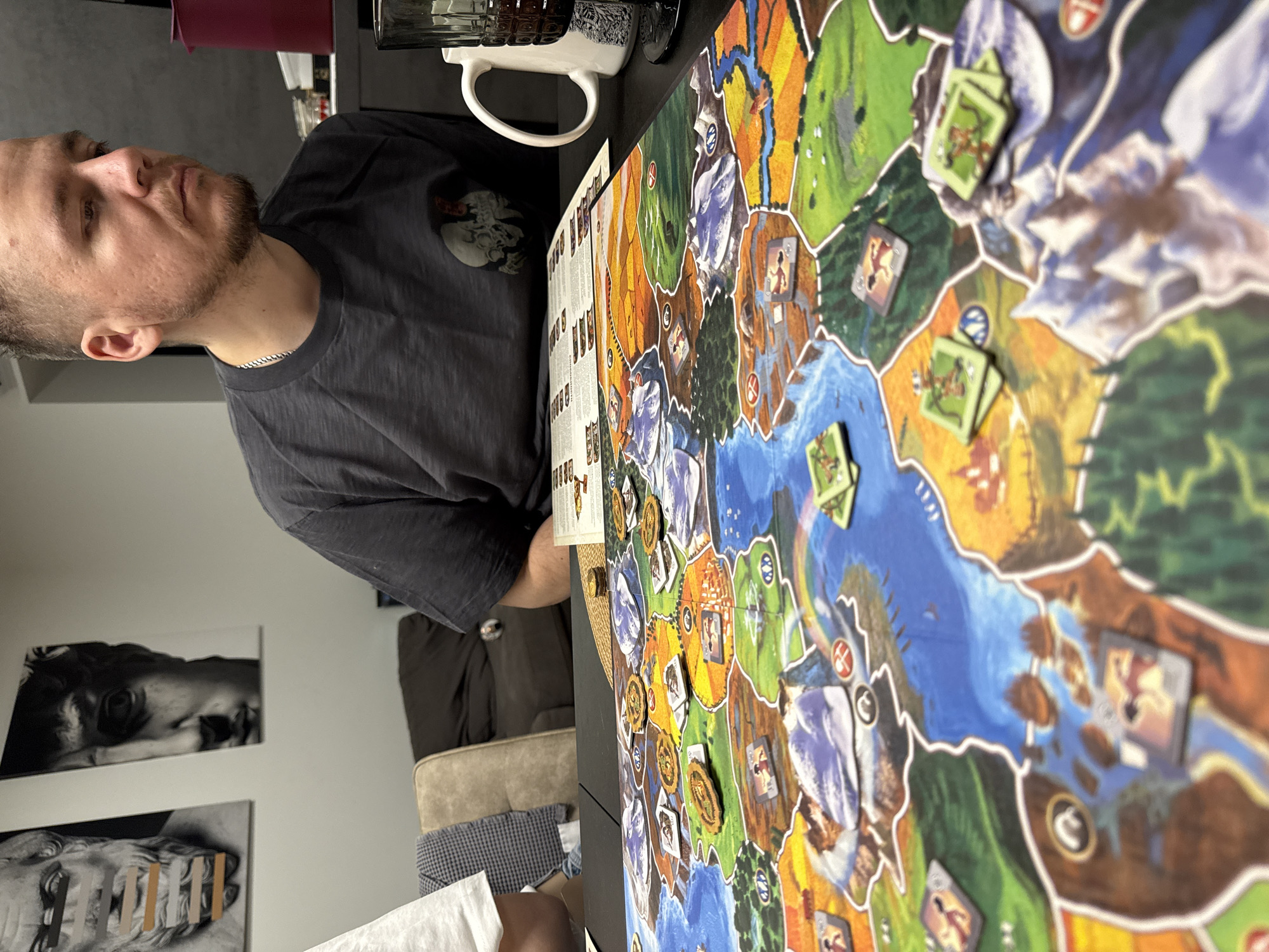 This media file captures an indoor scene in Krasnoyarsk, Russia, featuring a board game session. The primary subject is a man, seen from the chest up, with short hair and a beard, wearing a dark grey t-shirt adorned with a skull graphic. He is wearing a silver chain and appears focused, looking intently towards the left of the frame, engaged in the game.
The central object is a large, colorful board game laid out on a dark table. The game board features a stylized map with various terrain types including green land, blue rivers and lakes, and white/grey mountains. Numerous game components are visible on the board: small square tokens, some depicting archers and other human-like figures, and round, golden-colored tokens. A white ceramic mug with a black pattern and another dark mug are positioned on the table near the board. Adjacent to the game board is a white sheet of paper or card, likely a rulebook or player aid, containing unreadable text and graphics.
In the background, two black-and-white art prints are mounted on a light-colored wall. The top print shows a close-up of a classical sculpture's eye and forehead, while the bottom print features a similar classical bust with abstract vertical elements obscuring its eyes and mouth. A portion of a light brown upholstered sofa or armchair is also visible in the lower left background, suggesting a comfortable living space. The lighting appears to be artificial, indicating an indoor setting. The man's serious and engaged expression suggests deep concentration in the ongoing game. A slight glimpse of another person's arm or hand in the bottom right corner suggests multiple players are participating.