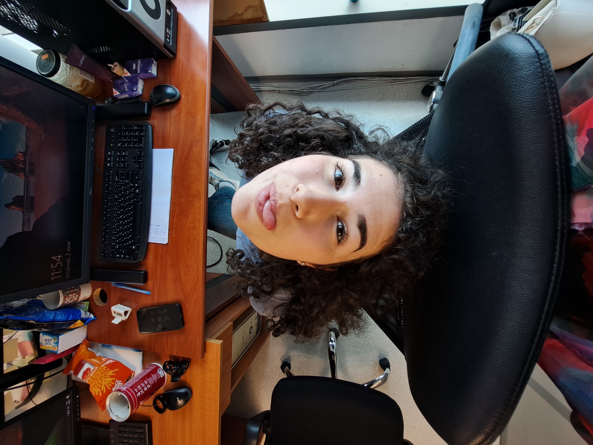 An overhead shot captures a young woman seated at a cluttered wooden desk in what appears to be an office or study space. The woman, with dark curly hair, looks directly up at the camera, playfully sticking out her tongue. She is wearing a grey top and blue jeans, with her legs and sneakers visible beneath the desk. The scene suggests a personal workspace in Sofia, Bulgaria, given the Cyrillic text visible.
The desk is densely populated with various items. On the left, a computer monitor displays "11:54 вторник, 2 юни" (11:54 Tuesday, June 2). A black keyboard and mouse are positioned next to it. Other desk items include a pen, a mobile phone, a bag of "POTATO STIX KETCHUP FLAV" snacks, a red Pringles-like can, a box labeled "VIEW CLEAR," and a mug. Towards the back of the desk, there are two boxes marked "SUPERSOFT," a tube of "euphoria" cream, and a jar containing a yellow substance. A computer tower or speaker system rests on a shelf above the desk.
The woman is seated in a black office chair with a mesh back. A second similar chair is partially visible in the bottom right corner of the frame. The flooring is a light-colored carpet or linoleum, and a wall with visible cables runs along the background. The overall impression is a casual and active personal workspace.