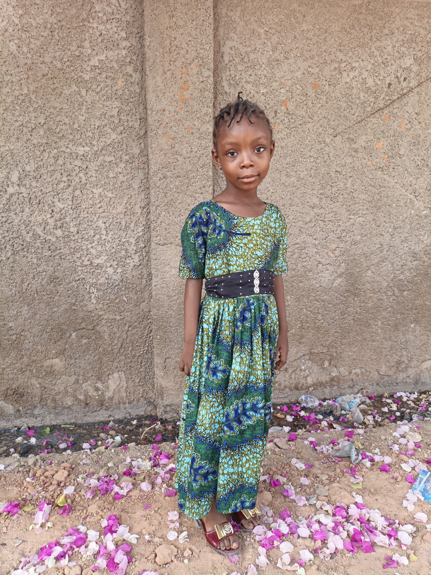 A young girl stands facing the camera in Gwagwarwa, Nigeria. She wears a floor-length, green and blue patterned dress with a black belt. The background is a rough, textured wall with a vertical seam. The ground is covered in red dirt and scattered with pink and white flower petals. There is trash visible near the base of the wall. The girl is wearing red sandals and has her hair braided.