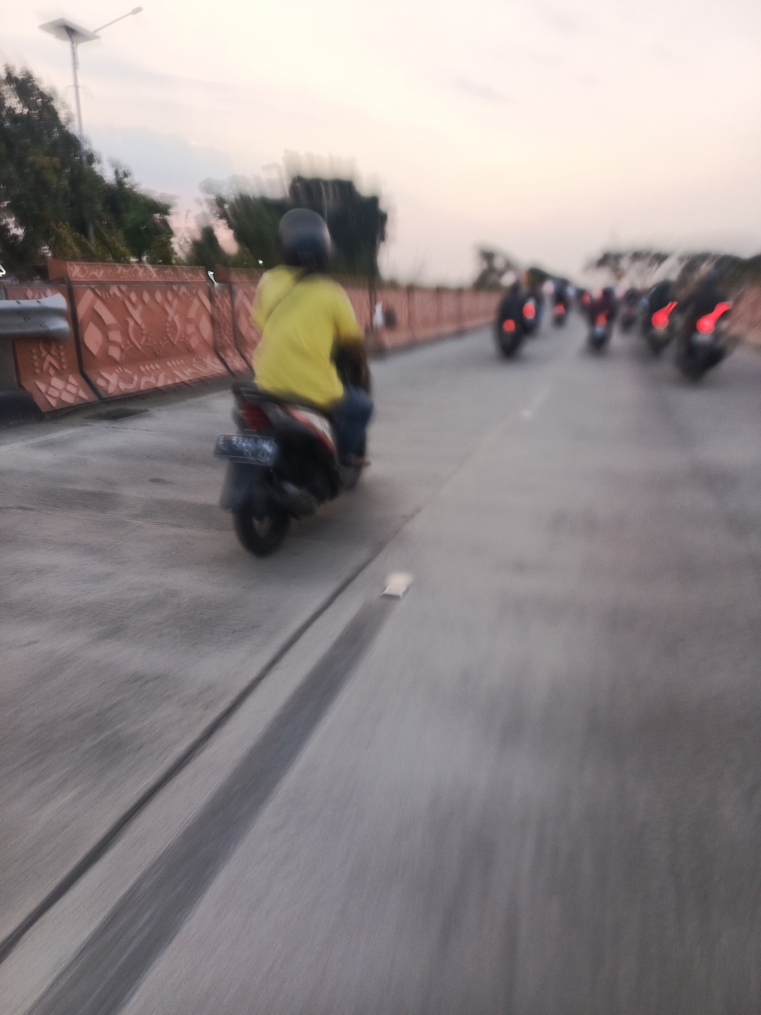 The image depicts a street scene at dusk in Sawotratap, Indonesia. A convoy of motorcycles is traveling on a paved road, with the lead motorcycle in focus. The rider on this motorcycle is wearing a yellow shirt and a dark helmet. The motorcycle itself is dark-colored with a license plate that is partially legible, showing "L 6124 BM". The road is flanked by a decorative orange barrier with a patterned design. In the background, trees and a street lamp are visible against a pale sky with hues of pink and orange, indicating the time of sunset. Other motorcycles are visible further down the road, their taillights illuminated. The overall impression is one of motion, with the blur in the image suggesting the motorcycles are in transit.