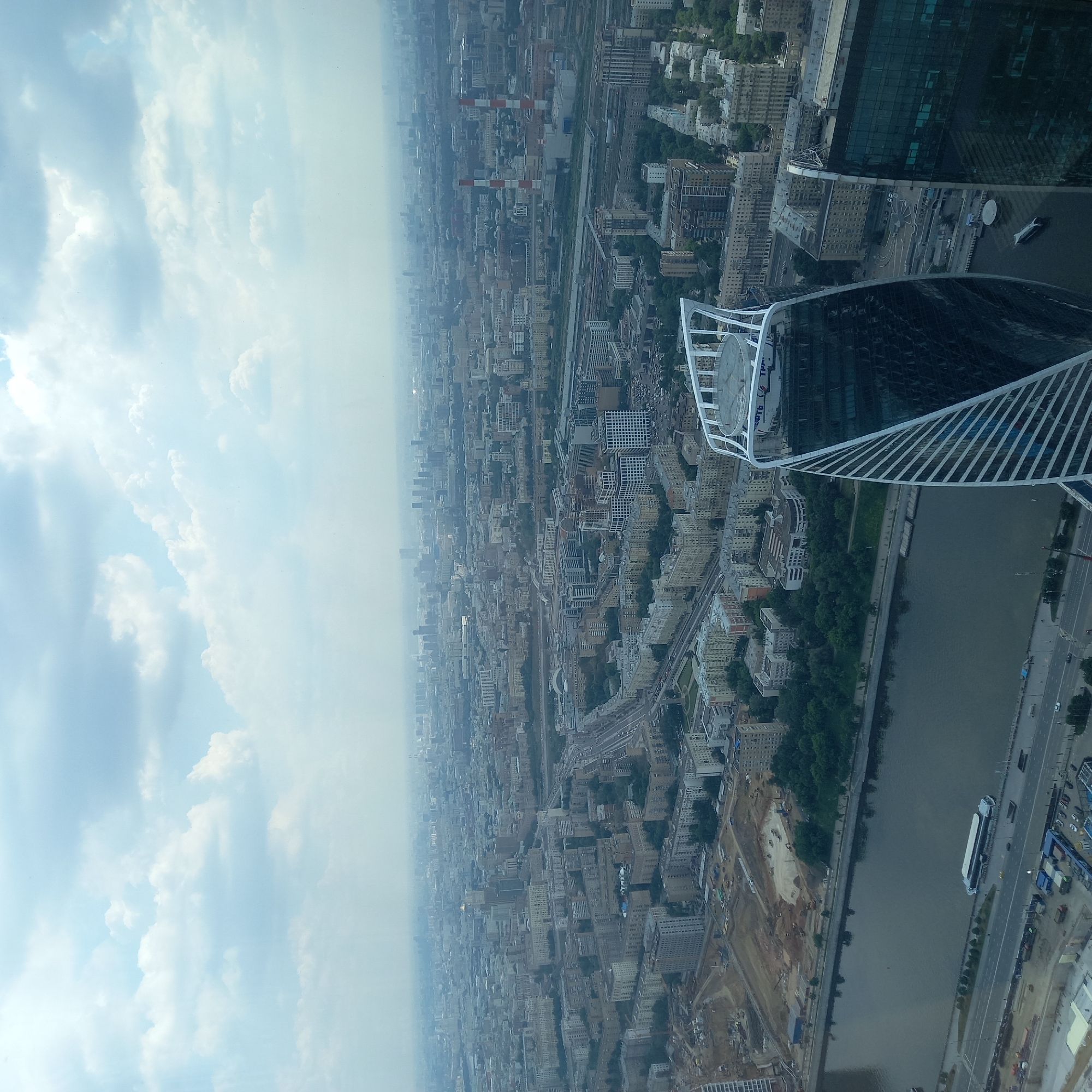 An aerial wide shot captures a dense urban landscape under a cloudy sky, dominated by the cityscape and a wide river. In the foreground, a distinctive, twisting skyscraper with a mirrored facade stands prominently, partially obscuring a neighboring, darker glass skyscraper. The river, a dark, reflective body of water, flows horizontally across the lower portion of the frame. On the river, a long, white passenger boat is moored near the bank.
Beyond the river, a sprawling metropolis unfolds, characterized by a vast expanse of buildings of varying heights and architectural styles, mostly in muted tones. Roads, highways, and what appear to be train tracks weave through the urban fabric. Construction sites, with exposed earth and machinery, are visible near the riverbank, indicating ongoing development. In the distance, the city stretches to the horizon, fading into a hazy, atmospheric perspective, with numerous smaller buildings and hints of industrial structures, including two tall, red and white striped smokestacks. The sky is filled with a thick layer of cumulus clouds, with diffused sunlight suggesting an overcast day. The overall impression is one of a large, busy city, a testament to human construction and activity against a natural backdrop of sky and water.