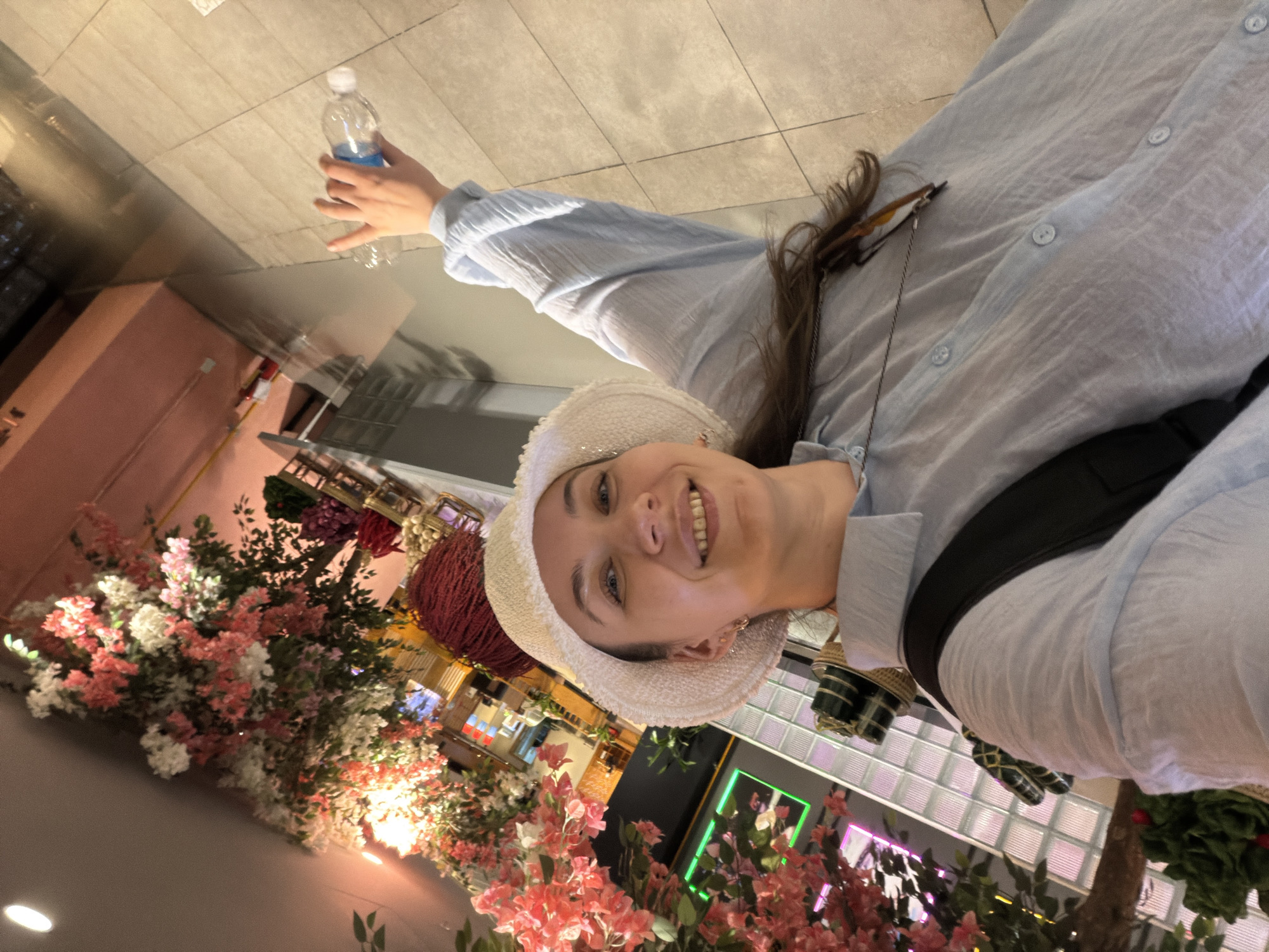 A woman in a light blue button-down shirt and a white hat is smiling and holding a water bottle in her raised right hand. She is in Nha Trang Ward, Vietnam, with an indoor floral arrangement consisting of pink and white flowers to her left and in the lower center of the image. There are shopfronts with green and pink neon signage visible in the background behind the floral arrangement. The floor is tiled.