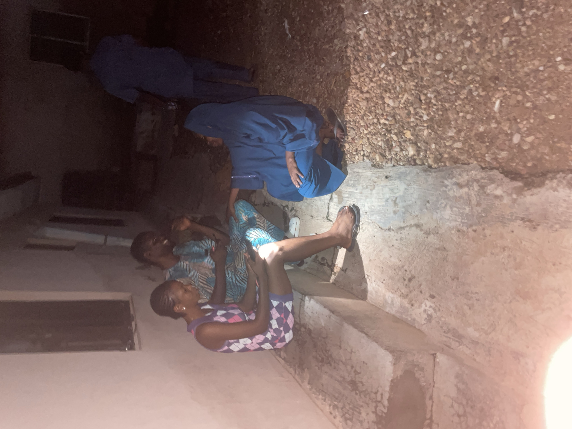 In Lokoja, Nigeria, at night, a group of individuals are gathered outdoors next to a building. Three people are clearly visible in the foreground. A woman in a sleeveless dress with a diamond pattern sits on a raised concrete ledge, looking at her phone. Beside her, a man in a patterned blue and green tunic also looks at his phone. To their right, a person wearing a blue hooded garment and matching pants is crouched down, facing away from the camera. Further in the background, another person in a blue shirt and pants stands, looking towards the group. The environment consists of a concrete walkway and a gravel-covered area. The lighting suggests it is evening or night.