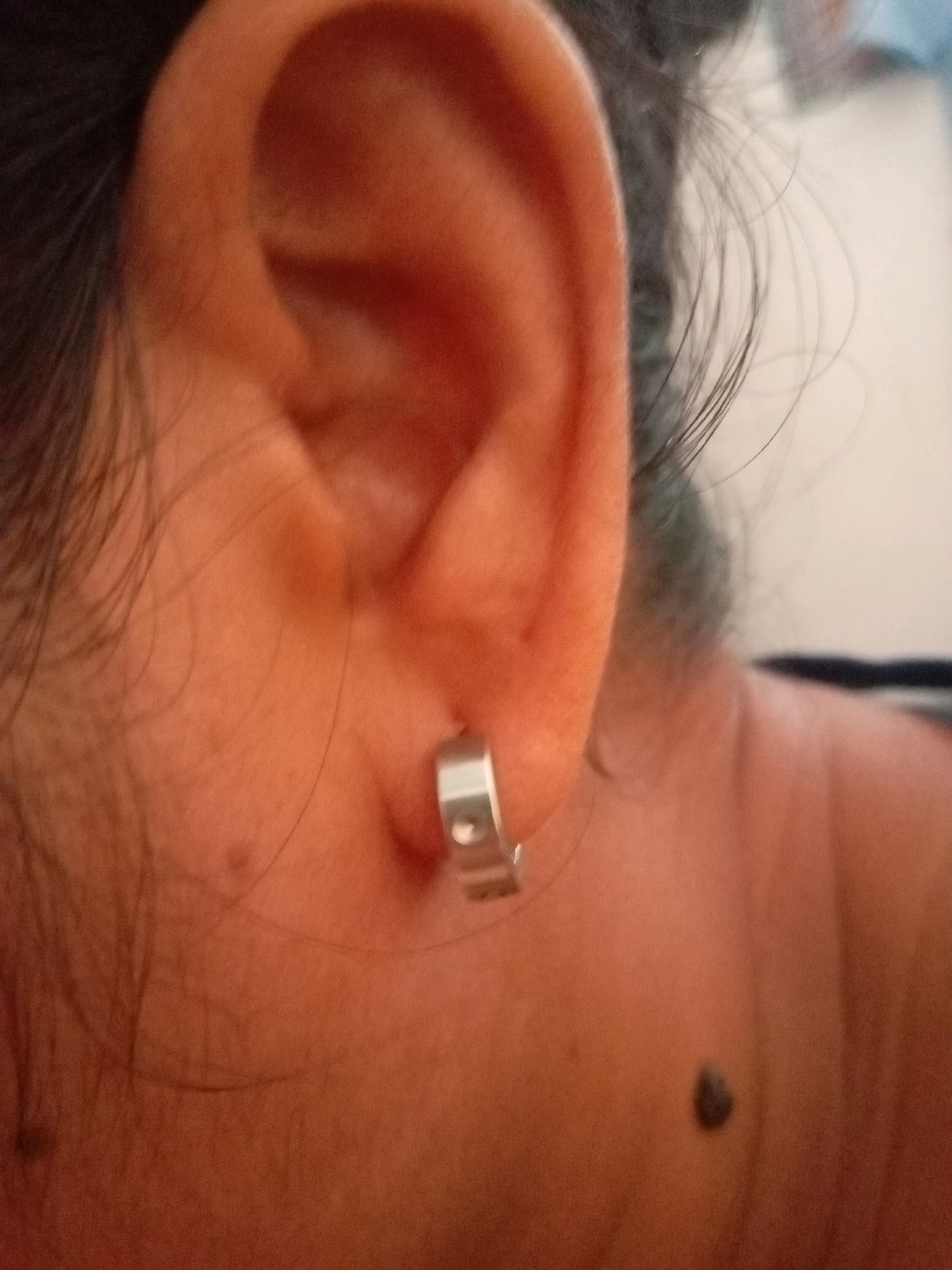 The image shows a close-up view of a person's ear and neck. A silver hoop earring is visible in the earlobe. The person has dark hair, some of which is visible framing the face. A mole is visible on the neck. The background is blurred and appears to be indoors. There is no visible indication of the location being Batam, Indonesia, or any specific city.
