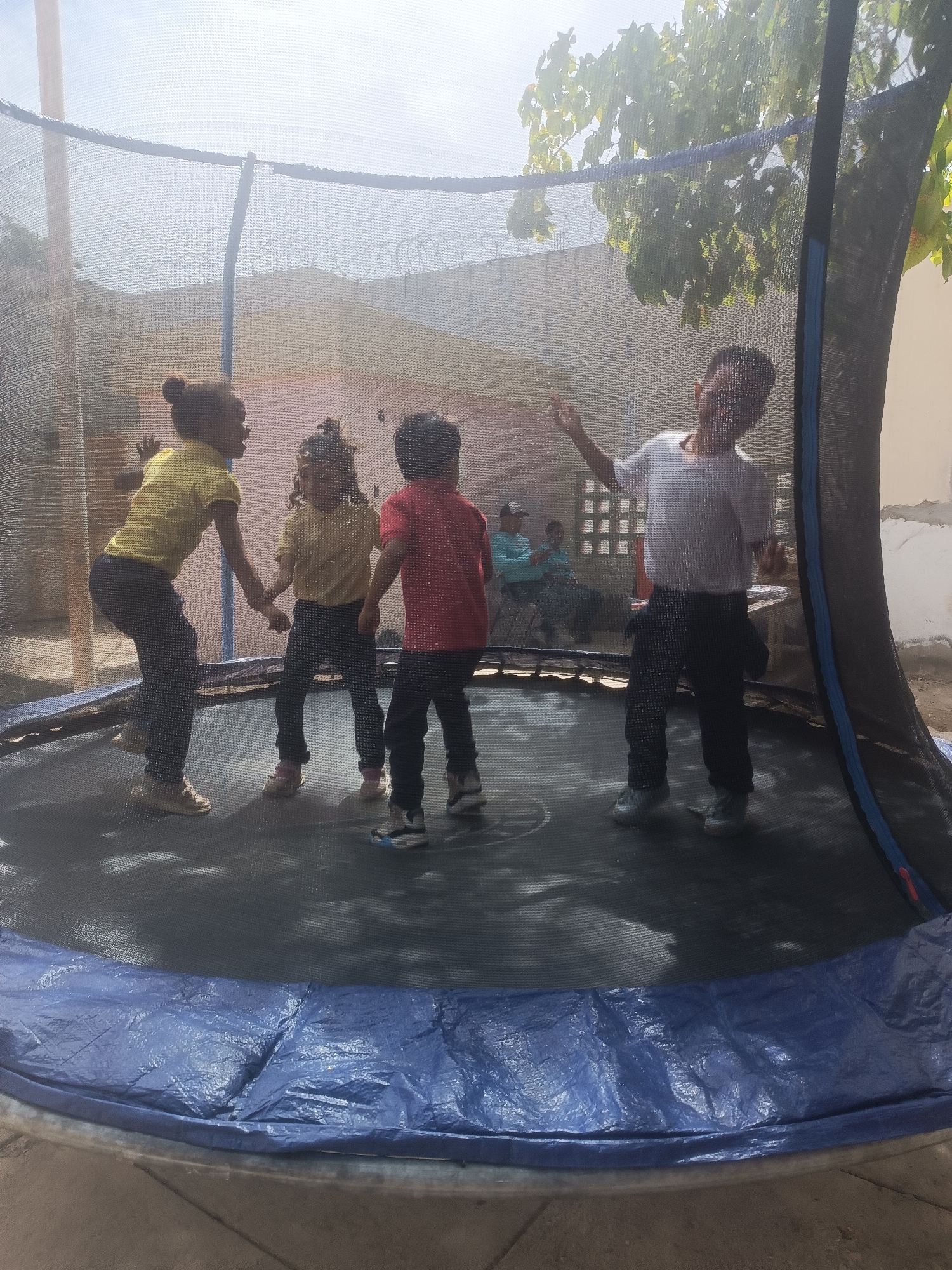 The image shows four children inside a trampoline enclosure. They are likely jumping and playing. Two adults sit on chairs in the background. The location appears to be outdoors, potentially a backyard, in the city of Porlamar, Venezuela. A wall with barbed wire is in the background.