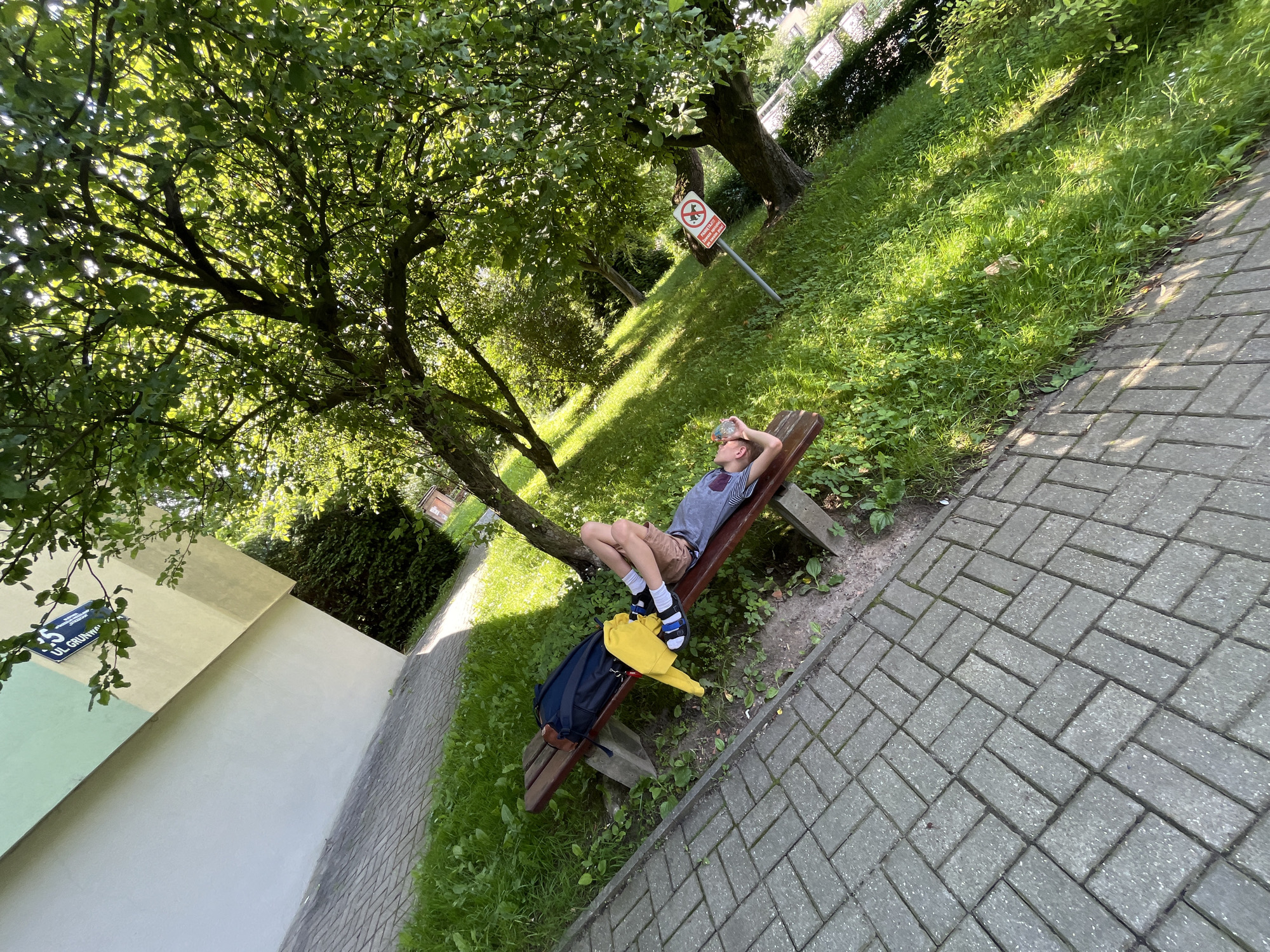 A young person lies on a park bench, their head propped up by their hands, eyes closed. They are wearing a blue t-shirt, brown shorts, and sandals. A yellow jacket and a blue backpack rest on the bench beside them. The bench is situated on a paved pathway, with a grassy area and a large tree to the right. A sign with a red circle and diagonal line, indicating a prohibition, is visible on a post in the grass. The background shows a building with a light yellow and green facade. The scene appears to be in a park or public green space.