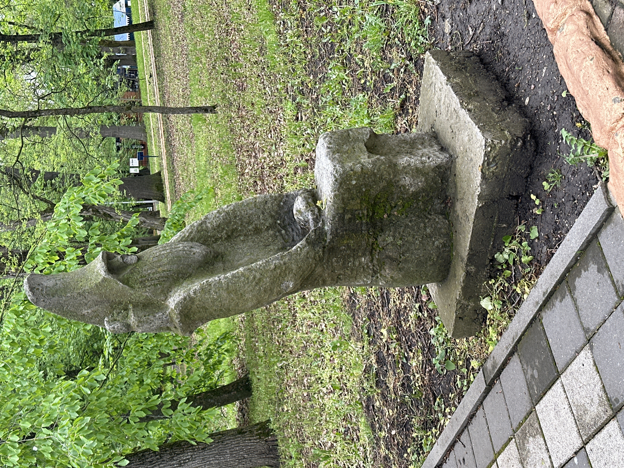 The image shows a stone sculpture situated on a concrete base in a grassy area next to a paved walkway. The sculpture appears to be an abstract representation of a bird-like figure. The surrounding environment includes trees and lawn. The location is Stavropol, Russia. No people are visible.