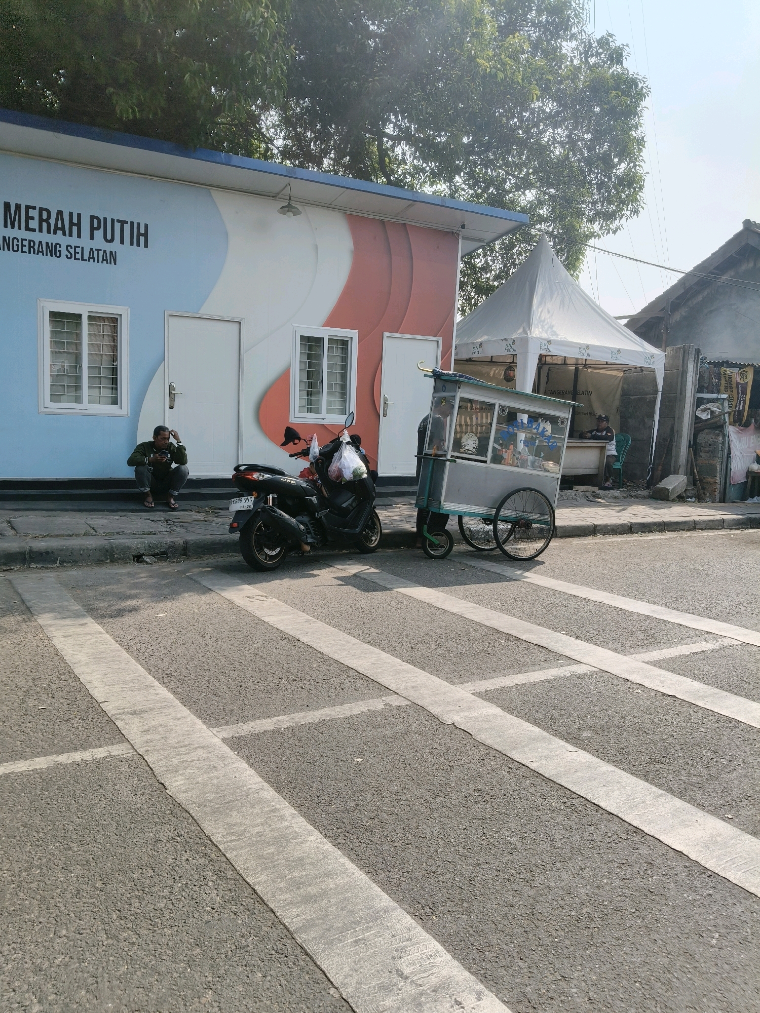 The image depicts a street scene in the city of South Tangerang, Indonesia, likely near a small commercial or administrative building. In the foreground, a paved road features pedestrian crossing markings. To the left, a one-story building with a blue and white facade is visible, bearing the text "MERAH PUTIH" and "TANGERANG SELATAN". Two windows with security bars and a white door are present on this structure. A man is seated on the curb in front of the building, facing towards the road and appearing to be using a mobile phone. Beside him, a black motorcycle is parked, with plastic bags hanging from its handlebars.
To the right of the building, a white tent structure is set up, possibly for a temporary stall or event. In front of the tent, a wheeled food cart is positioned. The cart appears to be made of metal and glass, with various items visible inside. Another individual is seated behind a counter within the tent area. The environment suggests a sunny day with trees and other buildings in the background.