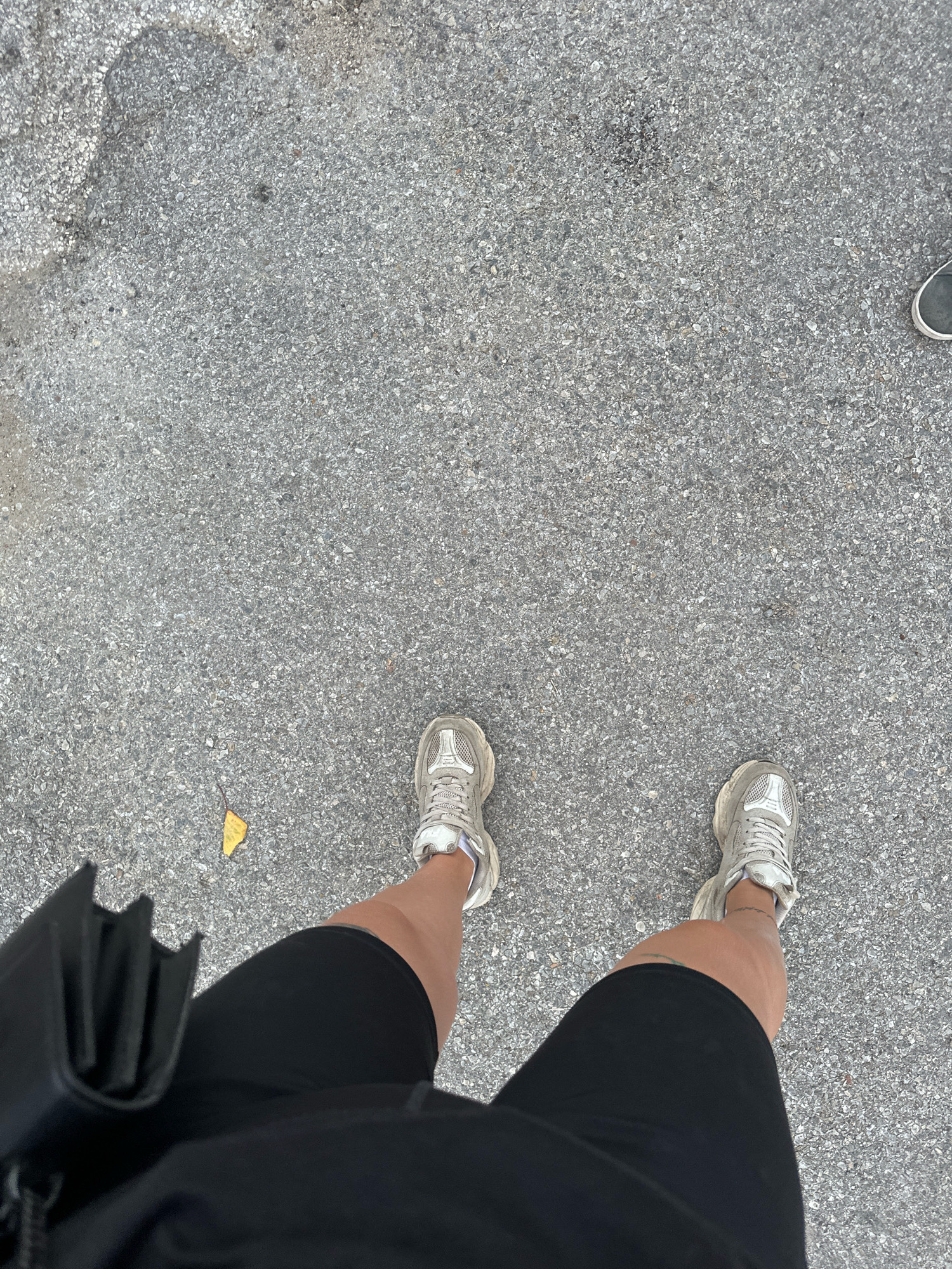 The image shows a person's legs and feet from a high-angle perspective, standing on an asphalt surface. The person is wearing black shorts and light-colored athletic shoes. A black bag is held in front of them, partially obscuring their left leg. A single yellow leaf is visible on the ground to the left of the person's feet. The asphalt appears to be rough and uneven, with small pebbles and aggregate visible. There is no indication of the location being Yavrovo, Bulgaria, or any specific city.