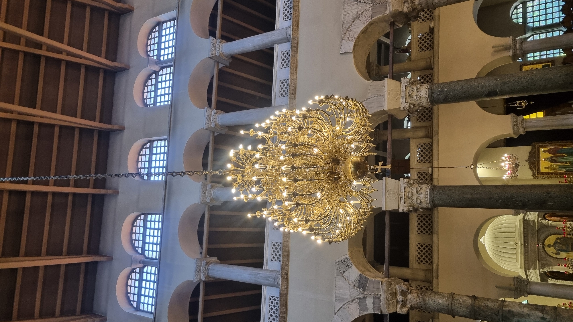 The image shows the interior of the Hagia Sophia Church in Thessaloniki, Greece. A large, ornate golden chandelier hangs from the ceiling. The chandelier has many branches with multiple light bulbs, illuminating the space. The ceiling is constructed with wooden beams. Above the chandelier, there are arched windows with latticed panes, letting in natural light. The church features a colonnade with marble columns and arched openings. In the background, there are glimpses of the church's dome and religious iconography, including a large framed icon with figures. The overall impression is of a grand and historic religious building, with a focus on the dramatic lighting provided by the central chandelier.