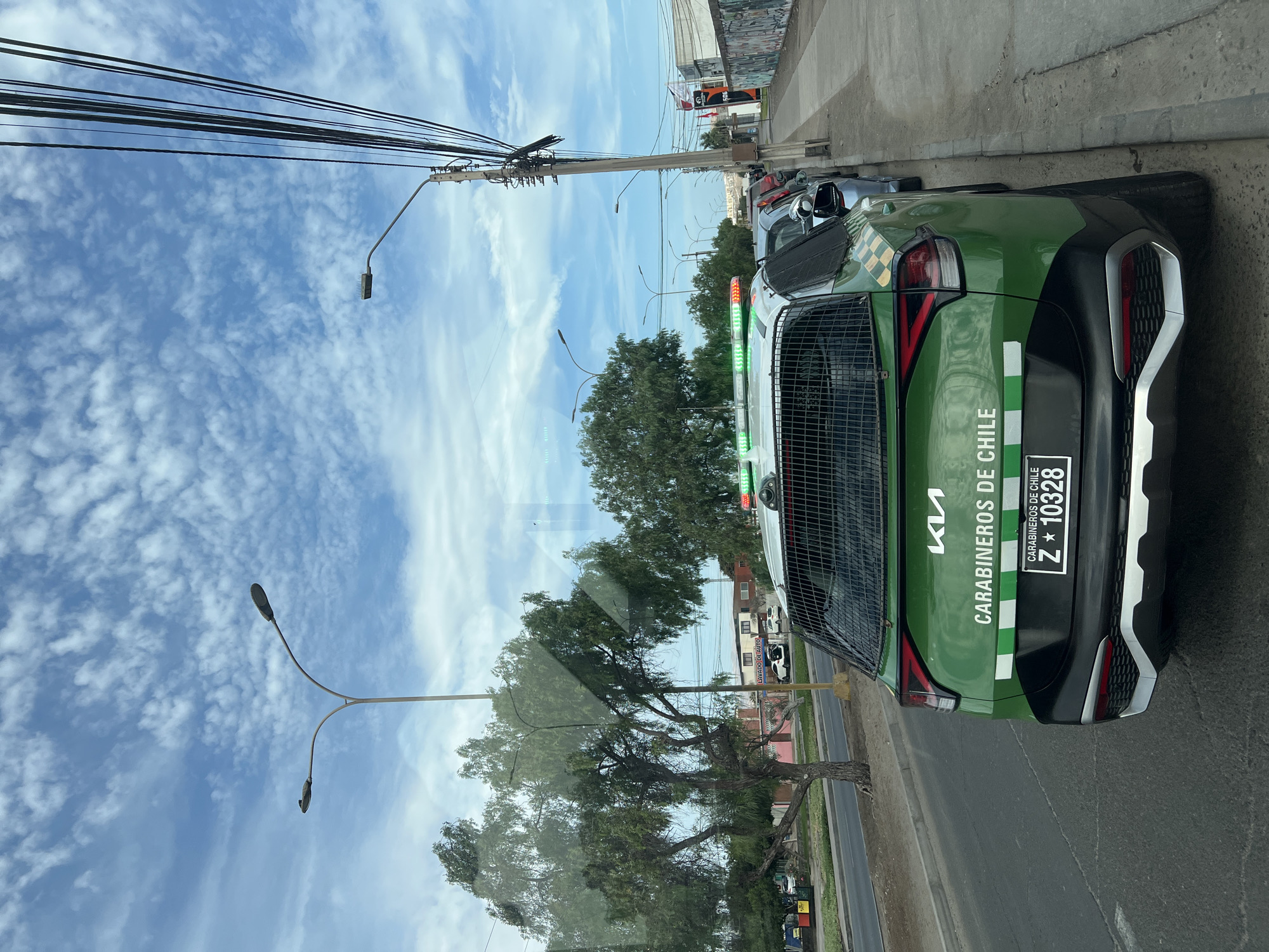 The image shows the rear of a green and black Kia Carabinero police vehicle parked on a street in Arica, Chile. The car has a metal cage over the back windows and a light bar on top. The license plate is visible and reads Z 10328. The vehicle is parked along a sidewalk next to a street with trees lining the side. Overhead, there are utility poles and wires. The sky is blue with scattered white clouds.