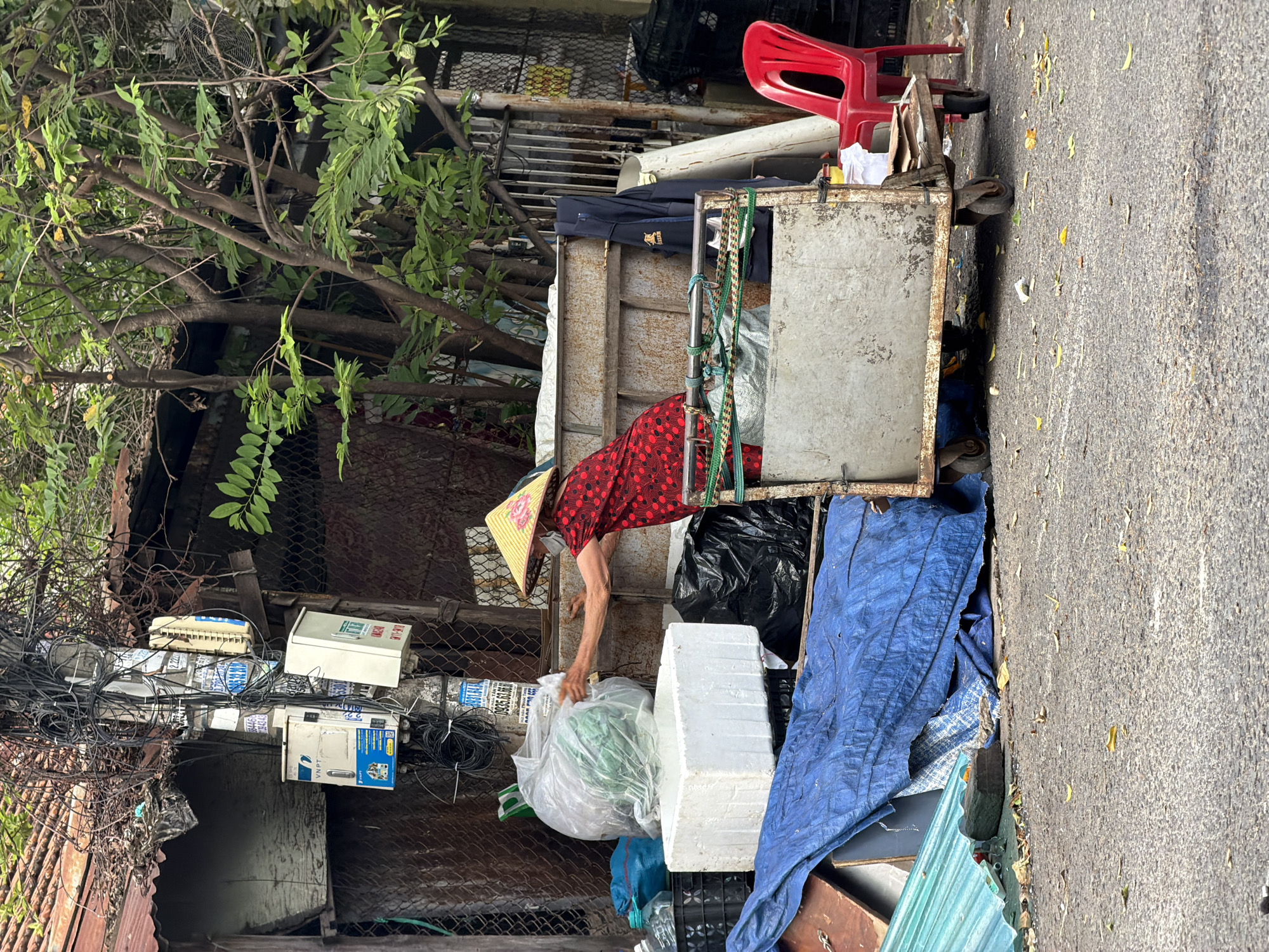 The image, viewed as if rotated 90 degrees counter-clockwise, depicts a daytime street scene in an urban or semi-urban environment, likely in Nha Trang Ward, Vietnam. The central figure is a woman, partially obscured by a conical hat (nón lá) adorned with a floral design and a light-colored face mask, wearing a red long-sleeved shirt with black polka dots. She is actively engaged, leaning into a large, rusty metal pushcart, and reaching into a clear plastic bag containing green leafy material, suggesting she is collecting or sorting items, possibly recyclables or discarded goods.
The pushcart is substantial, filled with various items, including several black garbage bags, white rectangular foam blocks, and a blue tarp. A red plastic chair is perched atop the cart near the right edge. Behind the woman and the cart, a dense arrangement of green tree branches and leaves is visible. To the left, an intricate network of tangled black wires, cables, and several utility boxes or meters are mounted on a pole-like structure, indicative of complex infrastructure. Text on one white box clearly reads "VNPT" (Vietnam Post and Telecommunications Group), while other blue labels show phrases like "CÁP ĐIỆN LỰC" (Electric Cable) and "2.2kW", with a partial phone number "0906..." also visible. Below this, a metal mesh fence runs across the frame.
The ground to the right of the cart appears to be a paved road or sidewalk, sparsely scattered with yellow leaves. The natural light suggests it is daytime. The overall impression is one of everyday activity in a bustling, somewhat cluttered street environment.