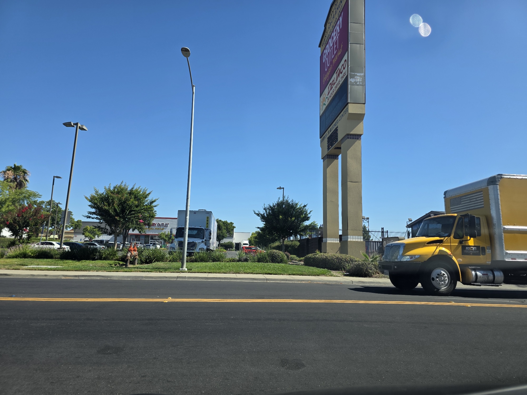 The image captures a commercial area in West Sacramento, United States of America, under a clear blue sky. A large, multi-columned sign for "GROCERY OUTLET BARGAIN MARKET" and "DELI TACO 24 HOUR DRIVE-THRU" is prominently displayed on the right side of the frame, rising high above the landscape. In the foreground, a paved road with double yellow lines runs horizontally across the bottom. To the right of the road, a bright yellow box truck with a silver trailer and an American flag decal on its rear is driving away from the viewer. The truck has the logo "ECF" visible on its side.
Behind the truck and across a grassy median, a parking lot is visible with several vehicles, including white sedans and a red sports car. A large white box truck is parked near a building. A fire hydrant, painted orange and red, stands near the grassy area. Further back, more trees and buildings, possibly storefronts, are seen. Several streetlights are scattered throughout the scene. A few individuals are visible in the distance, near the parking lot, seemingly interacting in the commercial environment. Lush green trees and bushes dot the landscape, interspersed with some flowering plants. The overall impression is a sunny day in a bustling commercial district.