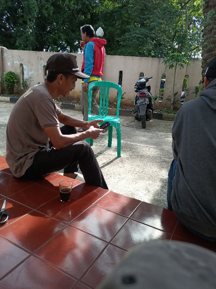 Three men are outdoors on a sunny day. One man sits on a tiled surface, looking at his phone, with a glass of dark liquid nearby. Another man stands in the background, wearing a red and blue jacket. A third man sits with his back to the camera, wearing a gray hoodie. A turquoise plastic chair stands behind the seated man with the phone. A motorcycle is parked near a concrete wall with rectangular cutouts. Trees and greenery are visible in the background.