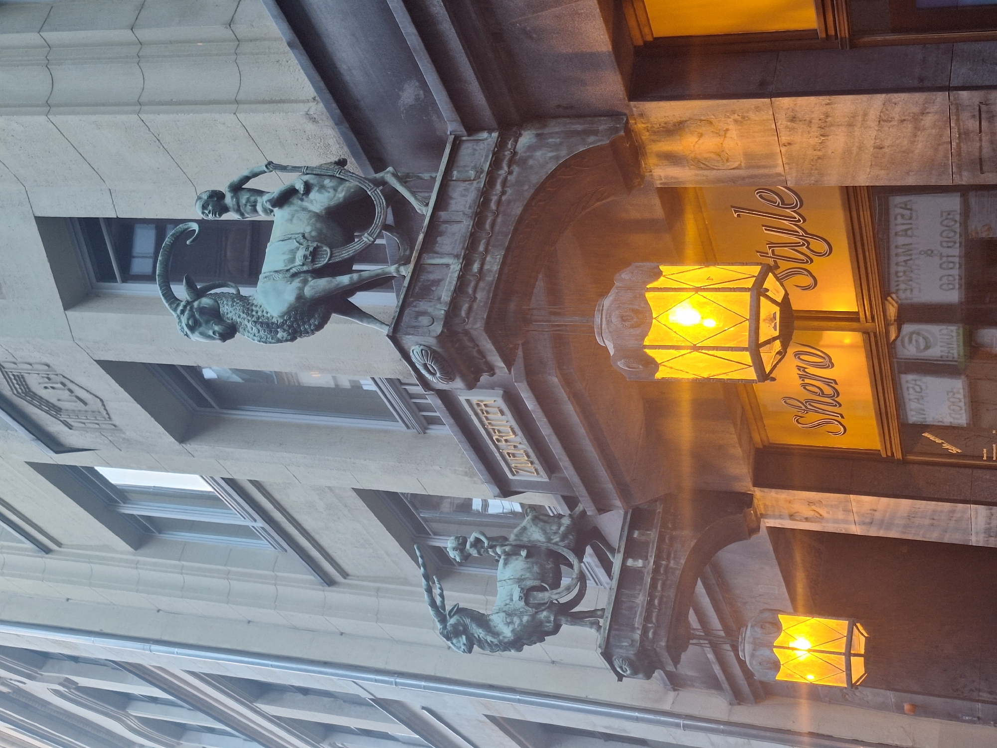 This is a medium-angle, eye-level, exterior shot of the historical building at Thomaskirchhof 12 in Leipzig, Germany. The facade of the building is made of light-colored stone blocks and features classical architectural elements.
Two ornate bronze sculptures of a goat being ridden by a cherubic child are mounted on cantilevered brackets projecting from the building. The sculptures are facing left and are positioned above arched entrances. The left sculpture appears to be slightly less detailed than the right one, but both depict the same subject.
Below the sculptures, on either side of the central entrance, are two large, hexagonal lanterns with yellow, illuminated glass panes. These lanterns hang from decorative metal brackets. The entrance itself has a sign that reads "Shero Style" in ornate, cursive lettering against a yellow background. Below this sign, a storefront window displays text, some of which is mirrored, suggesting it might be an "Asia Market" or similar establishment, with additional text visible such as "FOOD TO GO".
The building's upper floors are visible, showing rows of windows with light-colored frames. The architectural style is reminiscent of early 20th-century European design, with decorative molding and stonework. The overall impression is one of historical charm and artistic detail, characteristic of a well-preserved urban environment.