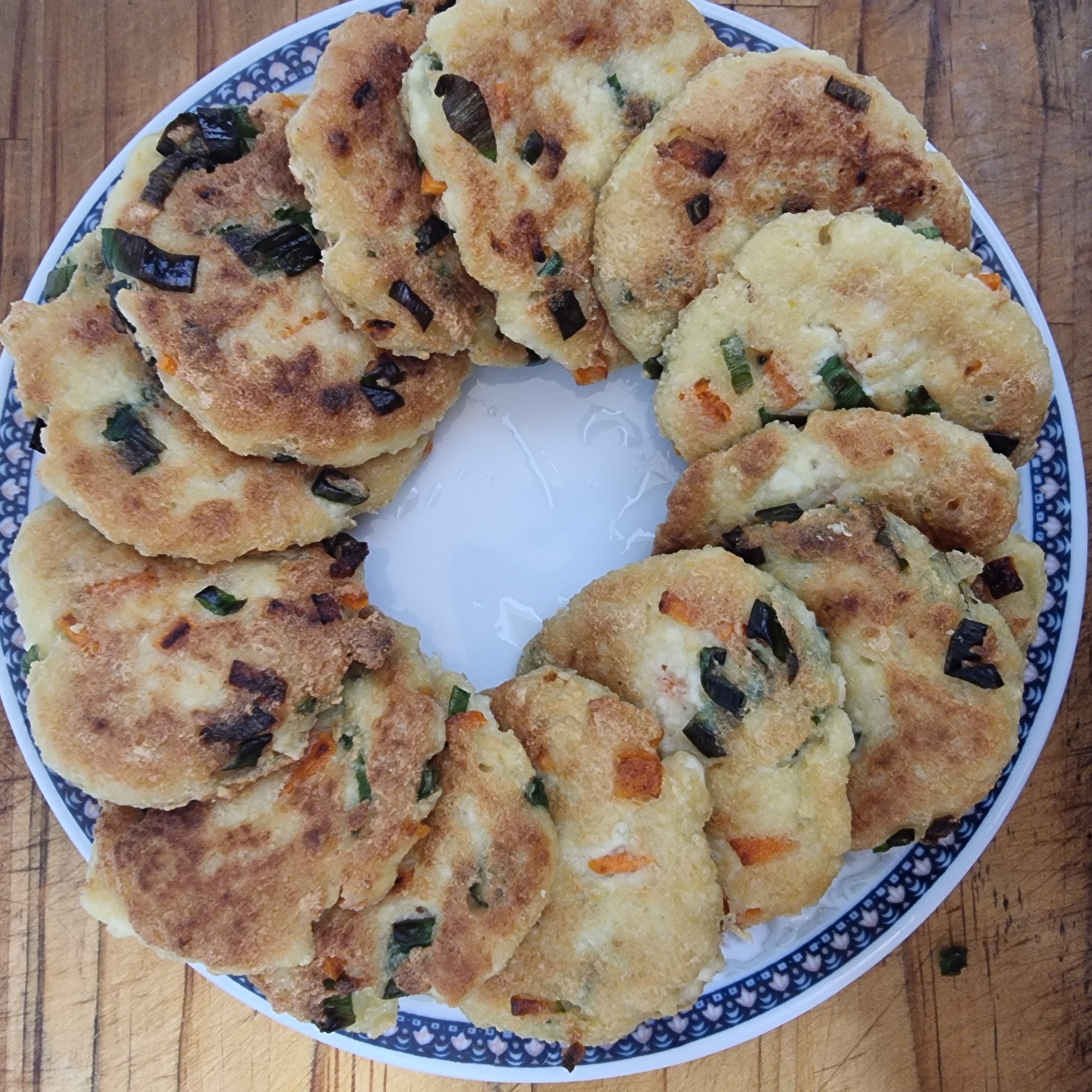 A plate of small, circular, pan-fried fritters is arranged in a circle on a wooden table. The fritters are golden brown and appear to be made from a batter mixed with chopped green onions and orange carrots. Some of the fritters have slightly darker, crispy edges. The plate itself is white with a decorative blue and white patterned rim. The wooden table has a visible grain. The image appears to have been taken in Dunnae-myeon, South Korea, as evidenced by the presence of what appears to be local cuisine served on a common style of dishware often found in that region. There are no people or animals visible in the image.