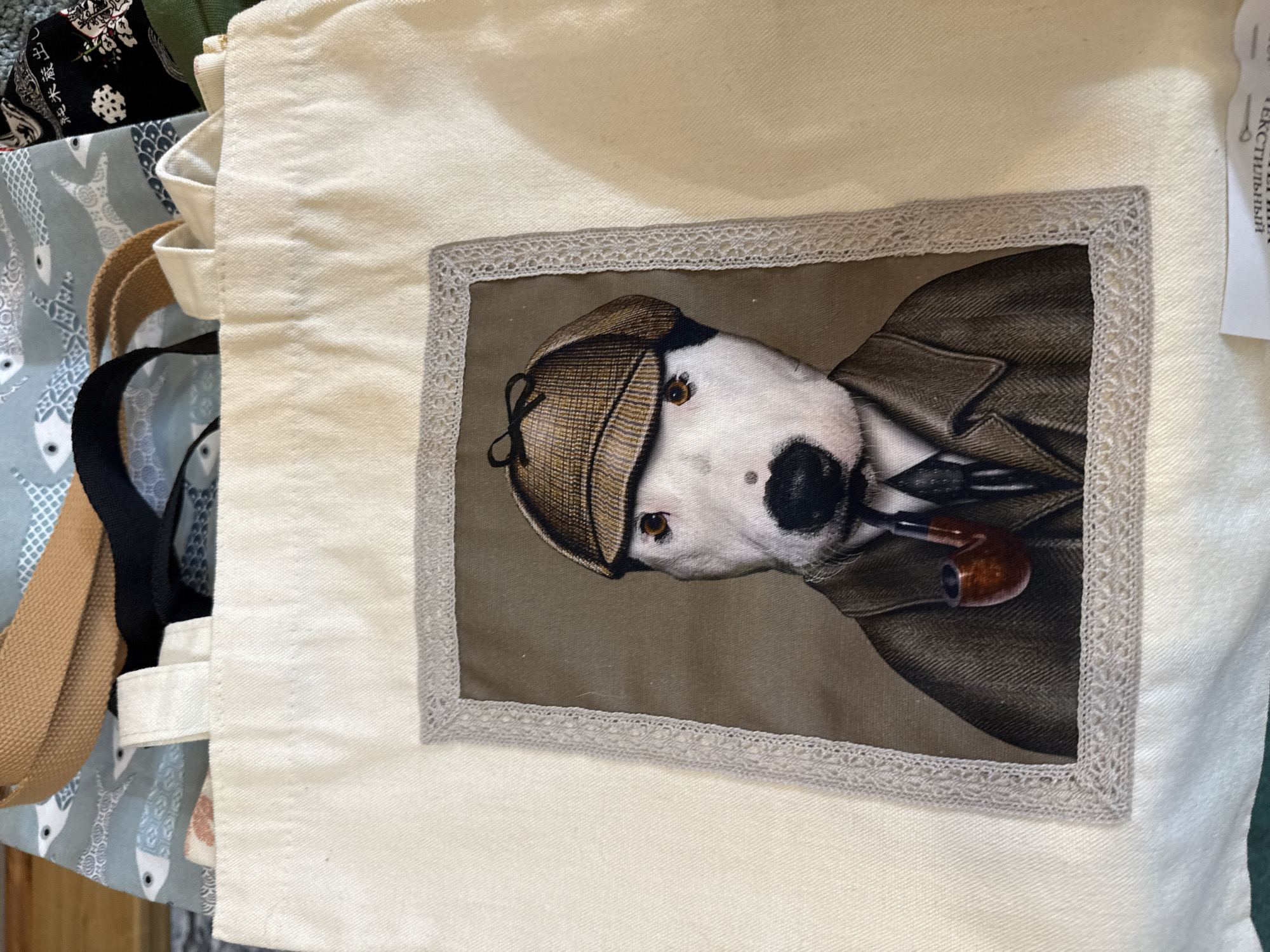 The image shows a close-up of a cream-colored canvas tote bag with a decorative patch. The patch features a detailed illustration of a white bull terrier wearing a tweed cap, a dark suit jacket, a tie, and holding a smoking pipe. The patch is framed with a delicate grey lace trim. The background of the tote bag is plain cream fabric, and other parts of the bag are visible, including straps and other fabrics peeking from behind, one with a pattern of small grey and white fish on a light blue background, and another black fabric with a floral pattern. The image is taken from a top-down perspective. There is no indication of the city of Khabarovsk, Russia, or any other specific location.