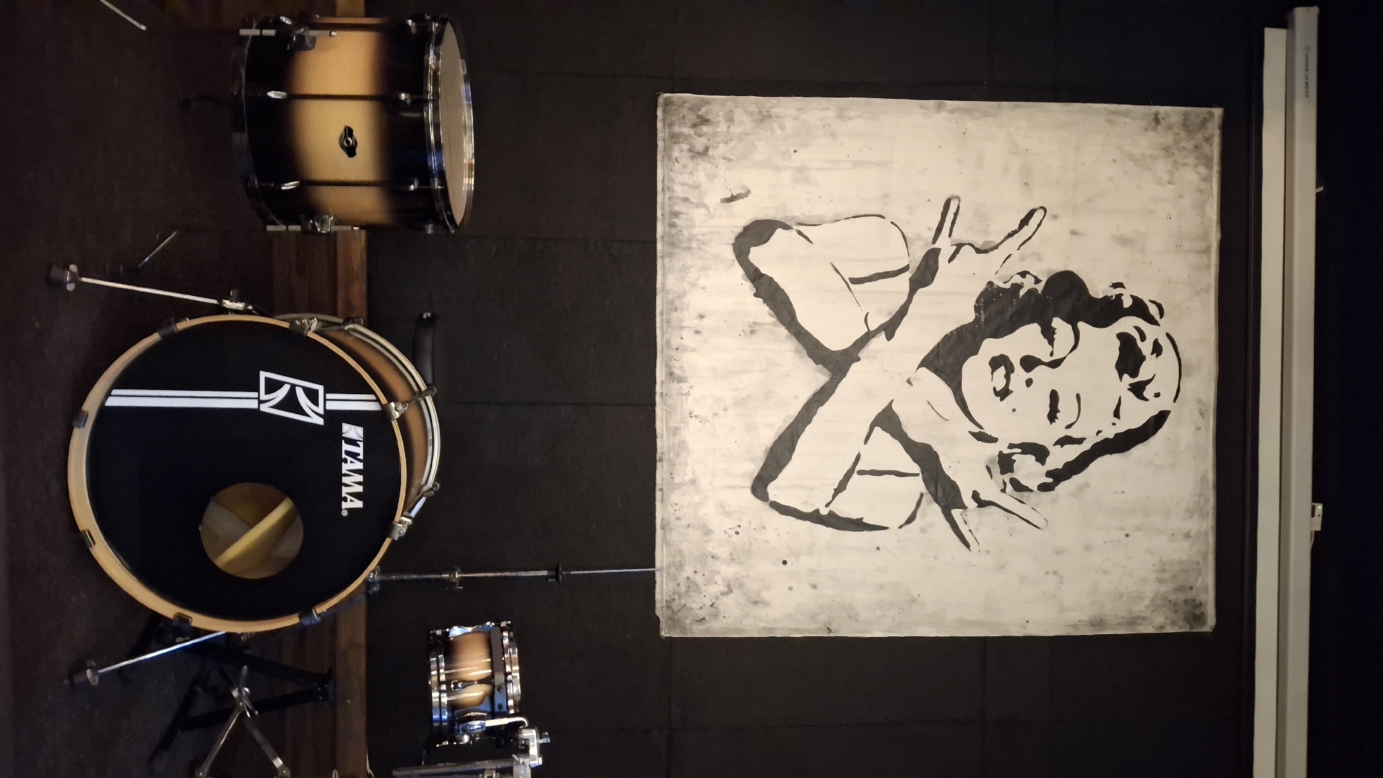 The image shows a large black and white stencil art of Marilyn Monroe with her arms crossed in front of her chest, hands held up in a peace sign, displayed on a wall. In the foreground, there are several drum set pieces: a snare drum on a stand, a bass drum with the Tama logo and a circular opening, and a tom-tom drum on a stand. The scene appears to be indoors, possibly a music studio or venue, with dark walls and flooring. A fluorescent light fixture is visible along the top of the wall.