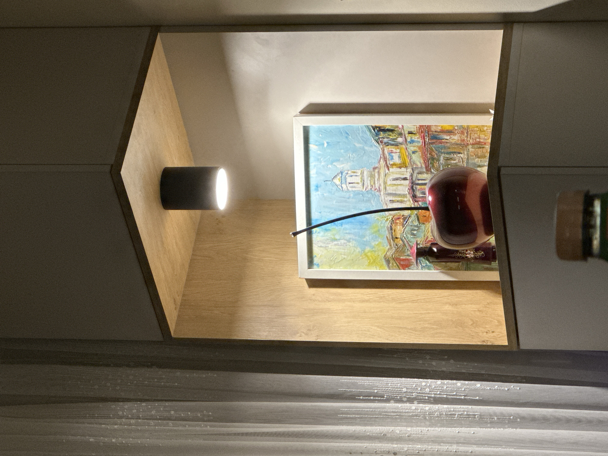 The image shows a framed painting of a building under a blue sky, possibly depicting a scene from Plovdiv, Bulgaria. A black cylindrical light fixture is mounted on the left interior wall of the alcove. To the right of the painting, there's a shiny, red cherry figurine and a partially visible bottle. The interior of the alcove is lined with light-colored wood. Below, a blurred surface with light patterns resembling water or textured material is visible. The photograph appears to be taken inside a building, perhaps a home or office space in Plovdiv.