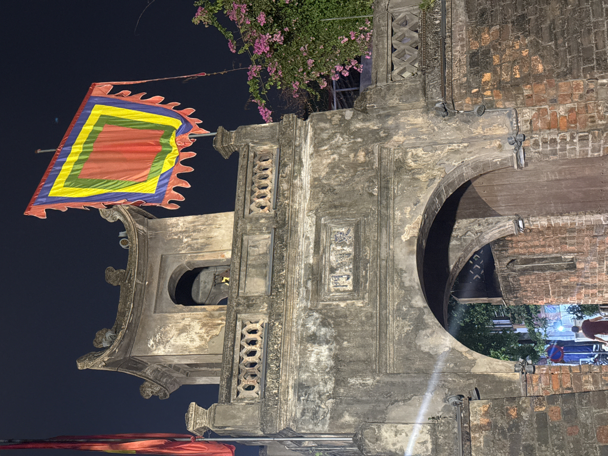 The image shows the imposing gate of the Temple of Literature (Văn Miếu-Quốc Tử Giám) in Hanoi, Vietnam at night. The gate is constructed of weathered stone and brick, with a traditional tiled roof and ornate carvings. A large, multi-colored flag, featuring concentric squares in red, yellow, green, and blue with a serrated edge, is prominently displayed on a pole to the right of the gate. To the right of the gate, a vibrant bougainvillea plant with pink flowers spills over a stone railing. The archway of the gate leads to a dimly lit inner courtyard, where a single individual in a maroon shirt is walking away from the camera. The sky is dark, indicating nighttime. The overall impression is one of historical grandeur and cultural significance.