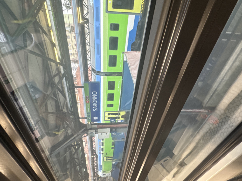 The image depicts a view from inside a train, looking out a window at a train station in Saronno, Italy. A prominent blue rectangular sign, centrally located, clearly displays "SARONNO" in white capital letters.
On an adjacent track to the right of the viewing train, another passenger train is stationary. This train features a distinct livery of bright green, blue, and white stripes/sections, with multiple dark-tinted windows and yellow-framed doors visible along its side. The front section of this train is visible on the far left.
The station platform area includes an overhead metallic support structure with visible girders and yellow-toned lighting fixtures. Below the adjacent train's windows, a blue tarp-like material covers an area of the platform, with a textured reddish-brown barrier or material visible further right. In the distant background, beyond the station structures, light-colored urban buildings are discernible under an overcast sky. No individuals are visible in the scene. The foreground of the image shows the interior of the viewing train's window frame and a reflection of a hexagonal pattern from inside the cabin.