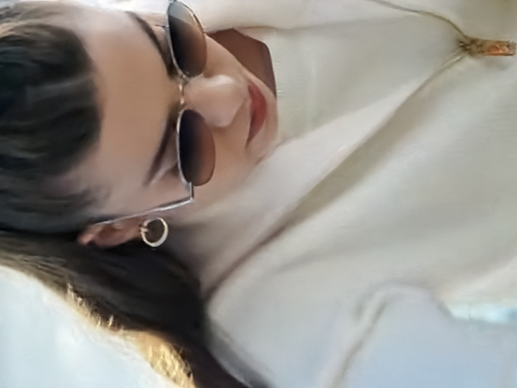 The image shows a close-up of a person wearing sunglasses and a white jacket. The person has dark hair pulled back and is wearing a circular earring and a pendant necklace. The background is blurry and appears to be an outdoor setting with a bright sky. There are no discernible landmarks or details to indicate the location is in the city of Kazan, Russia. The image does not contain any action or interaction between subjects.