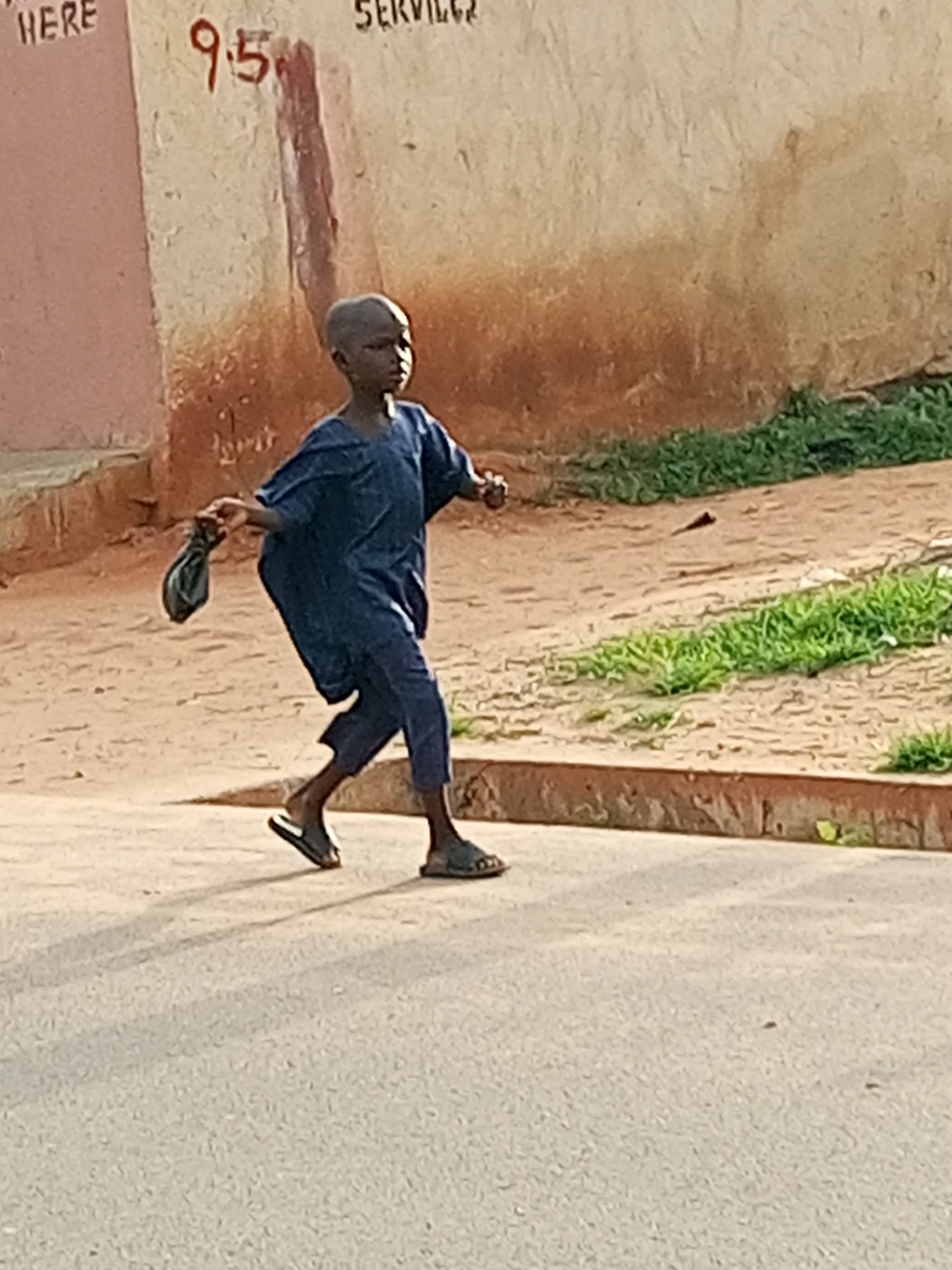 A young boy with a shaved head walks to the right on a paved street, carrying a dark cloth bag in his right hand. He wears a dark blue tunic and pants, and dark sandals. Behind him, a dirt path runs along the base of a textured, light-colored wall with red and brown staining. The wall has red lettering that reads "9.5" and possibly "SERVICES" above it. Green grass grows along the edge of the path. The scene is illuminated by bright sunlight, casting shadows on the street.