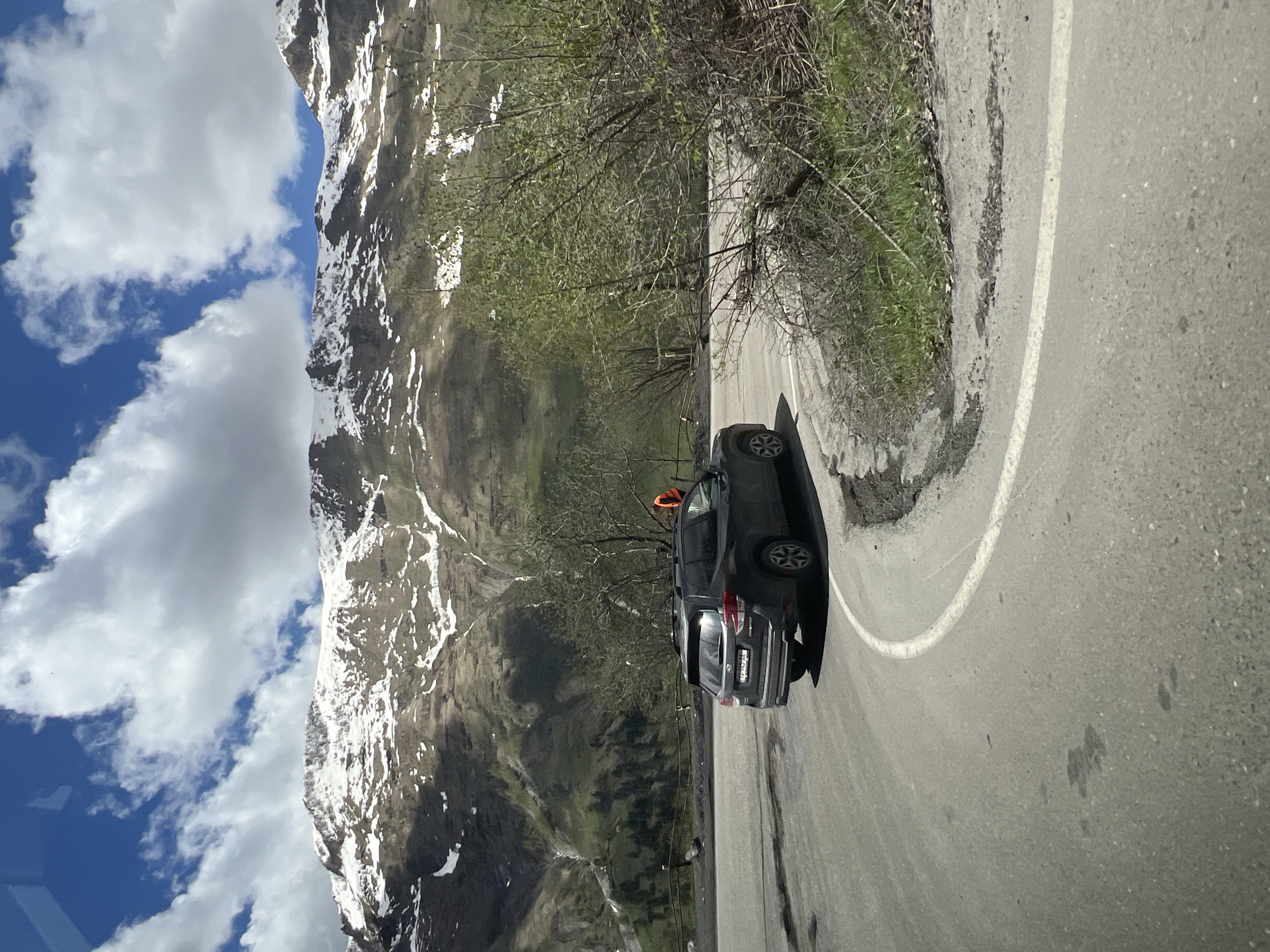 A car is parked on a winding road in Zemo Mleta, Georgia, with mountains visible in the background. The mountains have patches of snow. The sky is blue with scattered clouds. The road has a white line marking the edge. Some trees are visible along the roadside. There is a person in the car.
