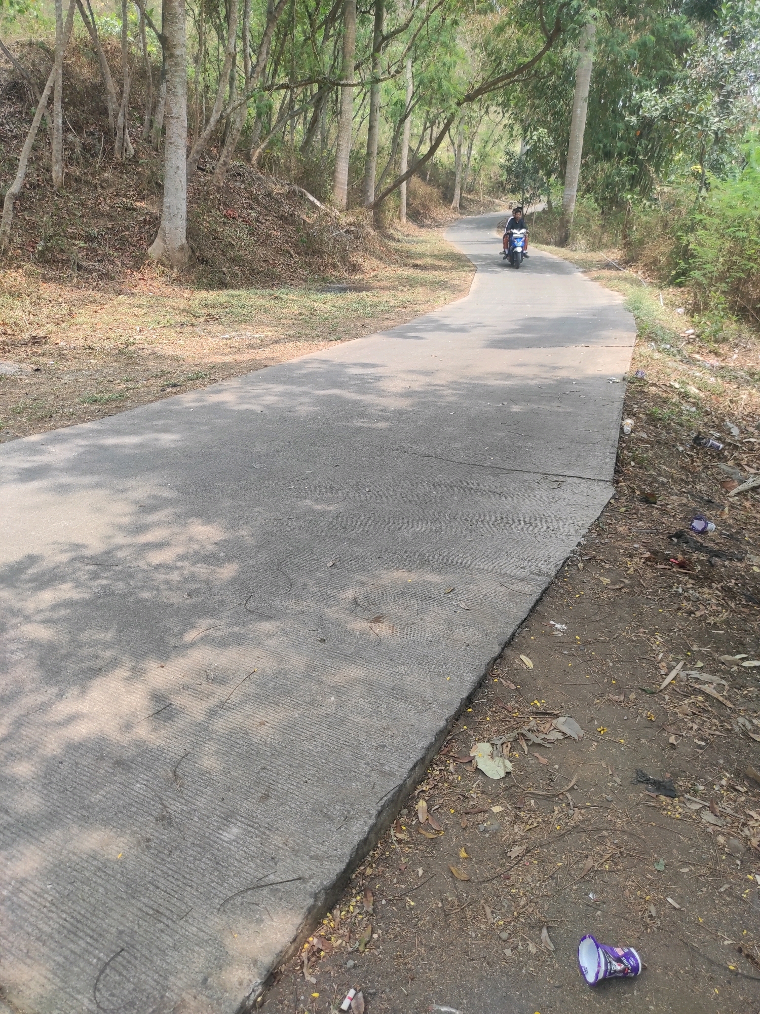 A paved, winding road curves through a wooded area. On the road, a person rides a blue and white scooter. The surrounding environment includes trees, dry leaves, and scattered litter, including plastic cups. The location is identified as Gunungmasigit, Indonesia.