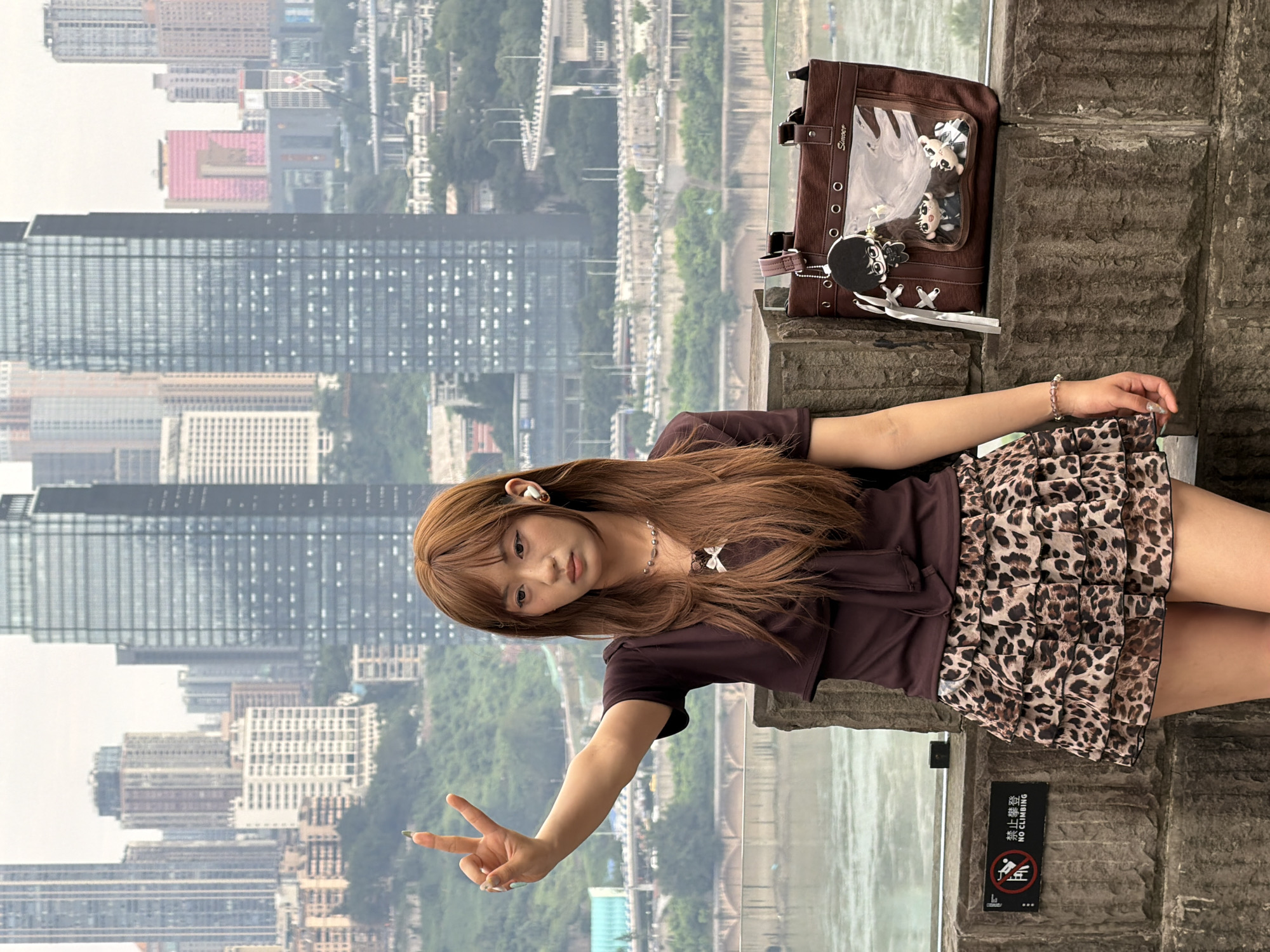In Yuzhong District, China, a young woman with long, brown hair stands on a stone ledge overlooking a city skyline. She is wearing a brown, short-sleeved shirt with a bow detail at the neckline and a leopard-print skirt. Her right arm is raised, and she is making a peace sign with her fingers. She has on white wireless earbuds. Beside her on the ledge is a brown tote bag with clear panels displaying several plush dolls. The background features numerous skyscrapers and a body of water. A "No Climbing" sign is visible on the stone ledge to her left.