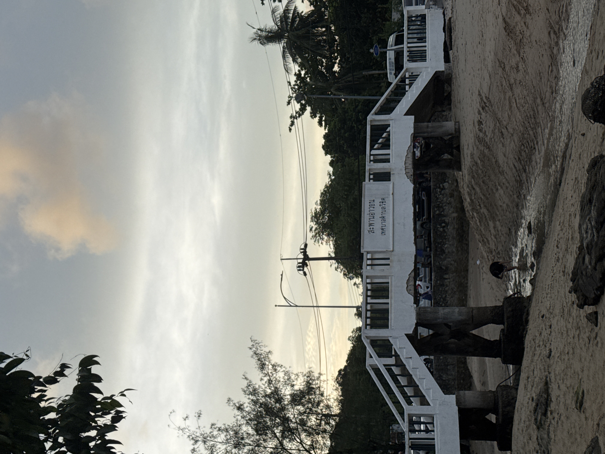 This image depicts a pedestrian bridge over a small stream or water channel on a sandy beach, likely in Wichit, Thailand. The sky is overcast with some patches of light-colored clouds, suggesting it might be late afternoon or early morning.
The bridge is white with black railings and has stairs leading up to it from both sides. The supports of the bridge are made of dark, weathered concrete pillars. On the bridge, there is a sign with Thai script.
In the foreground, the sandy beach is visible, with some rocks and debris scattered around. A person, wearing dark clothing, is standing in the shallow water of the stream, bending down as if examining something.
In the background, there are trees and vegetation, including palm trees, which are characteristic of tropical regions like Thailand. A road with some vehicles, including a white van, can be seen behind the bridge, suggesting it is near a populated area or a tourist spot. Power lines and a lamppost are also visible. The overall impression is a somewhat utilitarian bridge on a natural beach setting, with signs of human activity nearby.