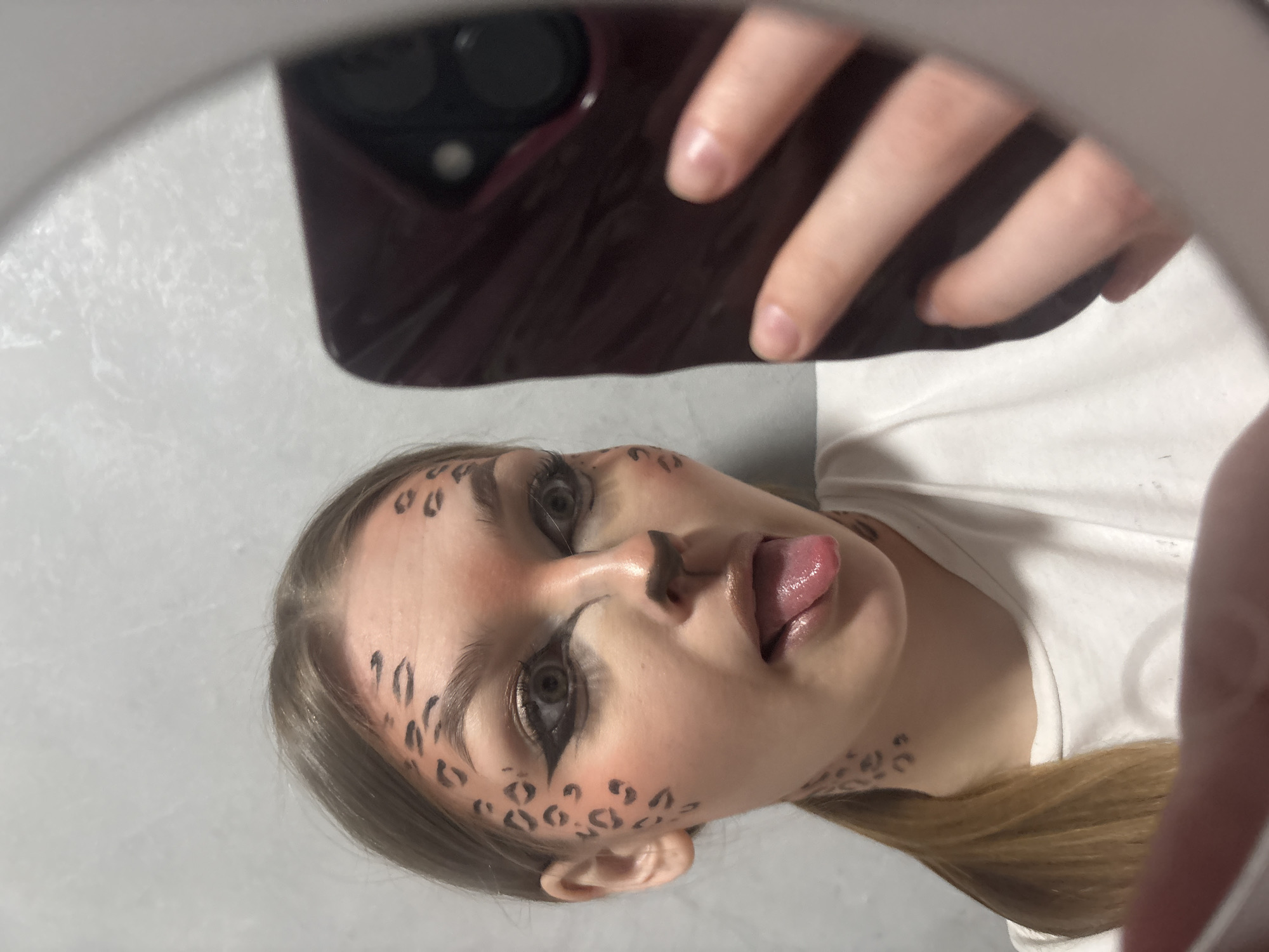 A person with painted cheetah spots on their face is visible in a mirrored selfie. The person is sticking their tongue out. They are holding a dark-colored smartphone, presumably to take the photo. The background shows a white surface and a portion of a white shirt. The person's hair is blonde. Location in the city of Chaikovsky, Russia is not discernible from the image.