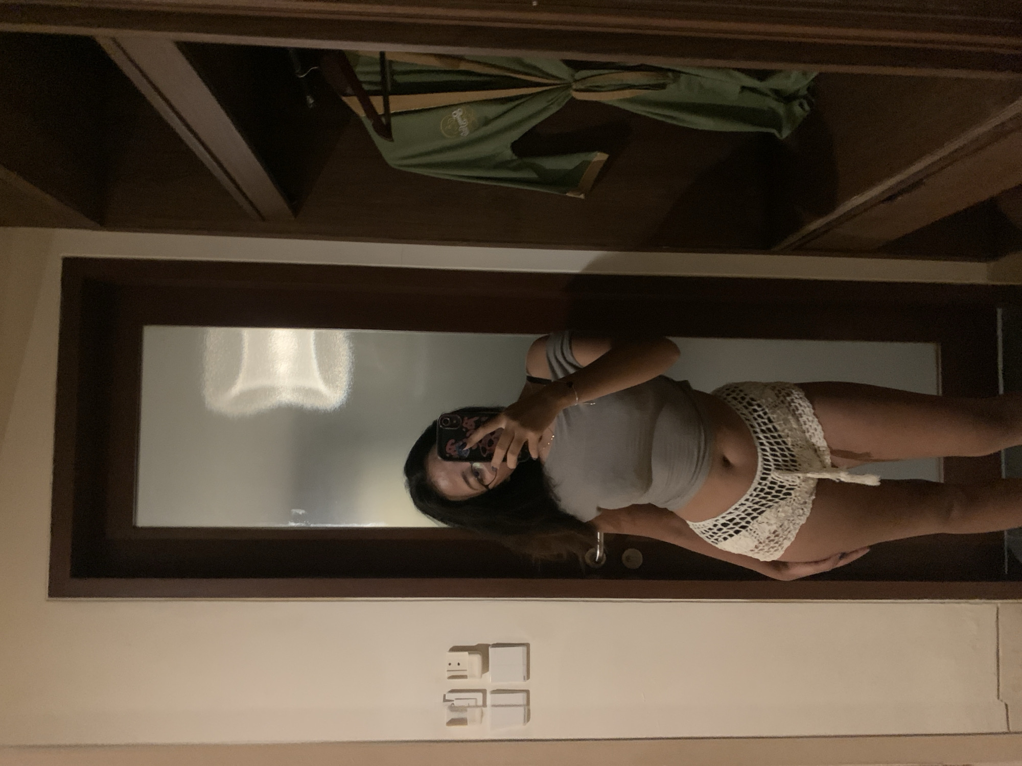 The image displays a mirror selfie taken indoors, likely in a hotel room or private accommodation within the Ubud District, Indonesia. A person, appearing as a young woman with long dark hair, is reflected in a large, vertical reflective surface, possibly a mirror or a glass door panel, framed in dark wood. She is wearing a grey crop top and white, intricately crocheted shorts.
Her left hand holds a smartphone with a decorative case, positioned to capture the selfie, partially obscuring her face. The activity depicted is the casual act of taking a self-portrait.
Above the main reflective surface, another reflection reveals the interior of a dark wooden wardrobe or cabinet. Inside, a green garment, possibly a robe with a light-colored, stylized logo, hangs on a hanger. The logo text is not clearly discernible. Below the reflective surface, a white wall is visible, featuring several rectangular light switches or electrical outlets. The indoor lighting suggests either daytime with interior lights on or an evening/night setting. No specific emotions are prominently displayed.