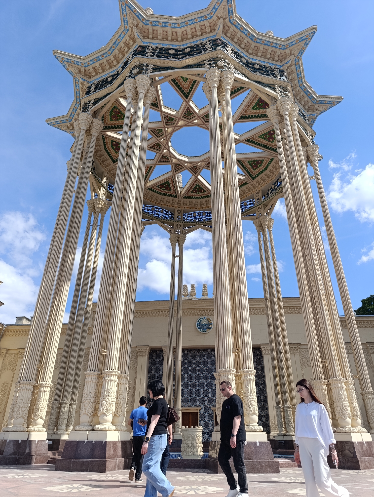The image shows the Friendship of Peoples monument in Moscow, Russia. The monument, a large, ornate structure with a star-shaped roof supported by numerous columns, is bathed in sunlight. The sky above is a clear blue with scattered white clouds. Several people are walking in the foreground, including a woman in a black t-shirt and jeans, a man in a black t-shirt and black pants, and another woman in a white top and white pants. The ground in front of the monument is paved with decorative tiles. The overall scene depicts a sunny day at a prominent landmark in Moscow.