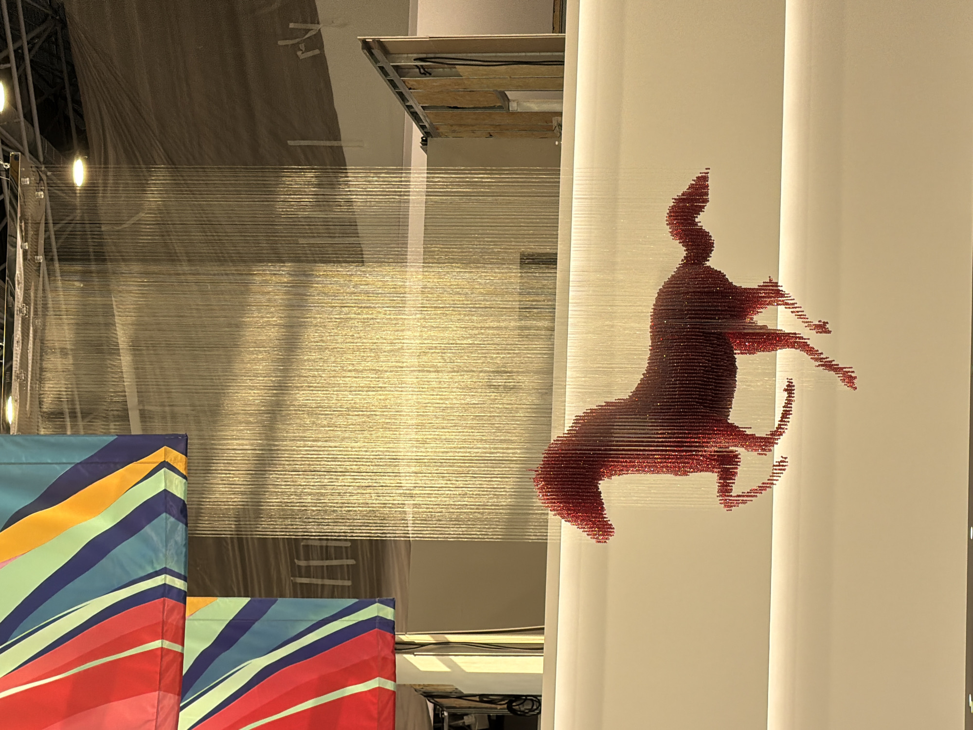 In an indoor exhibition space with a muted color palette, a red, beaded sculpture of a horse in mid-gallop is suspended in front of a shimmering, vertical curtain of thin, gold-colored threads. The sculpture's form is outlined by the individual beads, giving it a pixelated appearance. Above the horse and the gold curtain, two large banners with bold, diagonal stripes in red, blue, green, and yellow hang from the ceiling. The background reveals the structural elements of the space, including exposed wall framing and ceiling fixtures. The lighting is warm and appears to be artificial, casting shadows and highlighting the texture of the gold thread curtain.