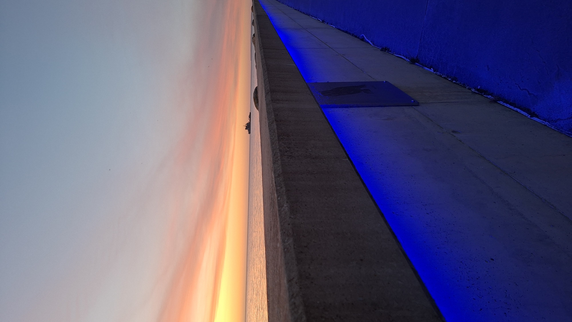 The image captures a sunset over a body of water, likely the ocean, as seen from a concrete walkway. The sky displays a gradient of colors, transitioning from a soft yellow and orange near the horizon to a pale blue overhead, with wispy clouds streaking across. A lone ship is visible on the water in the distance. The foreground features a thick, grey concrete wall or barrier running parallel to the walkway. A vibrant blue LED light strip is installed along the lower edge of this wall, illuminating the adjacent walkway. The walkway itself appears to be paved with a dark blue material. One small, square plaque is affixed to the concrete wall. The overall scene suggests an urban waterfront setting at dusk.