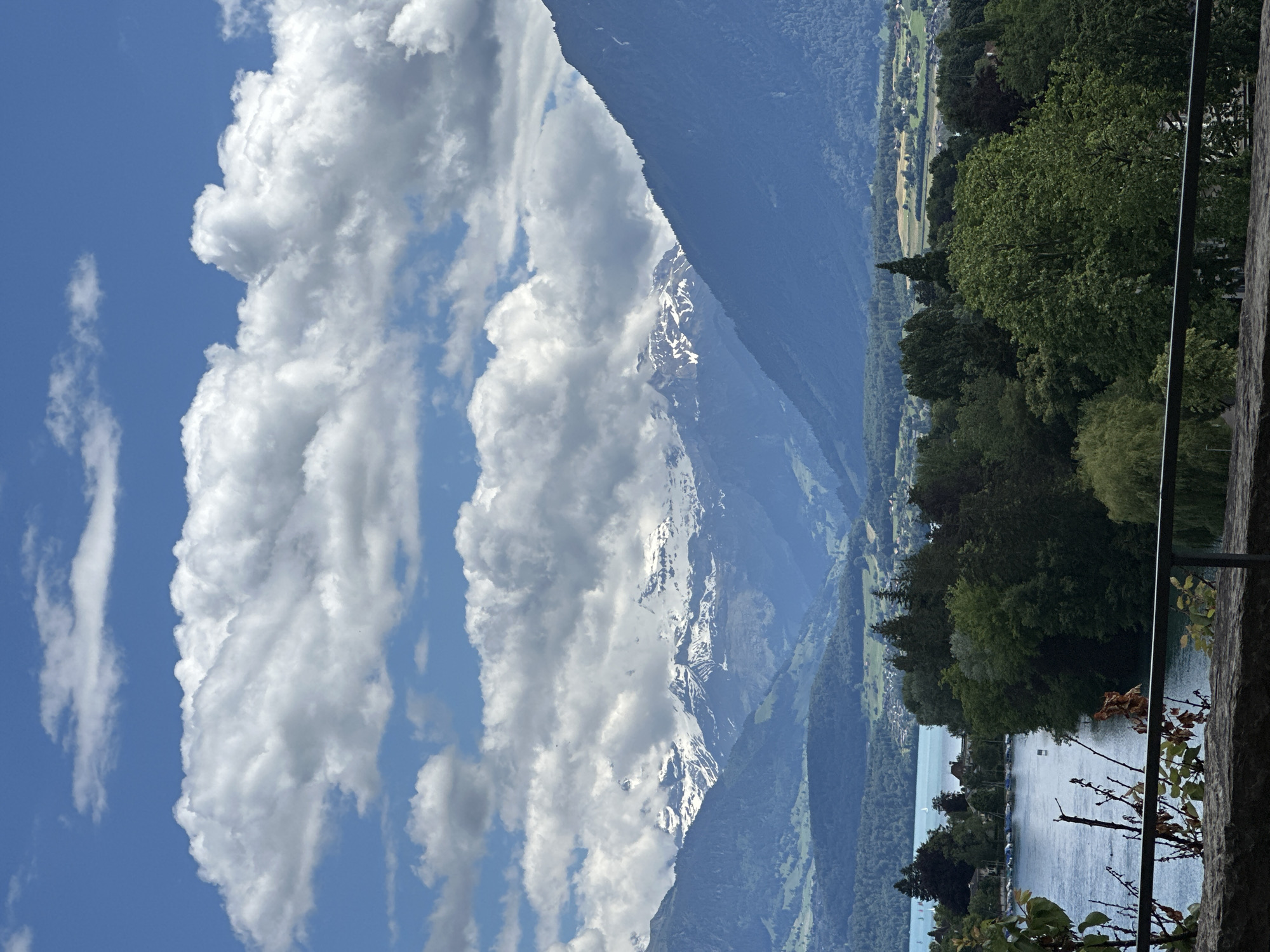 A scenic view of the city of Thun, Switzerland, is presented, featuring a vast expanse of the sky adorned with fluffy white clouds against a brilliant blue backdrop. Below, majestic snow-capped mountains rise, their peaks partially obscured by dramatic cloud formations. The midground is a tapestry of lush green trees and rolling hills, interspersed with the tranquil waters of a lake. Small boats dot the lake's surface, and houses are nestled amongst the foliage, hinting at the vibrant life within the city. In the foreground, a stone wall and railing offer a vantage point for this breathtaking panorama.