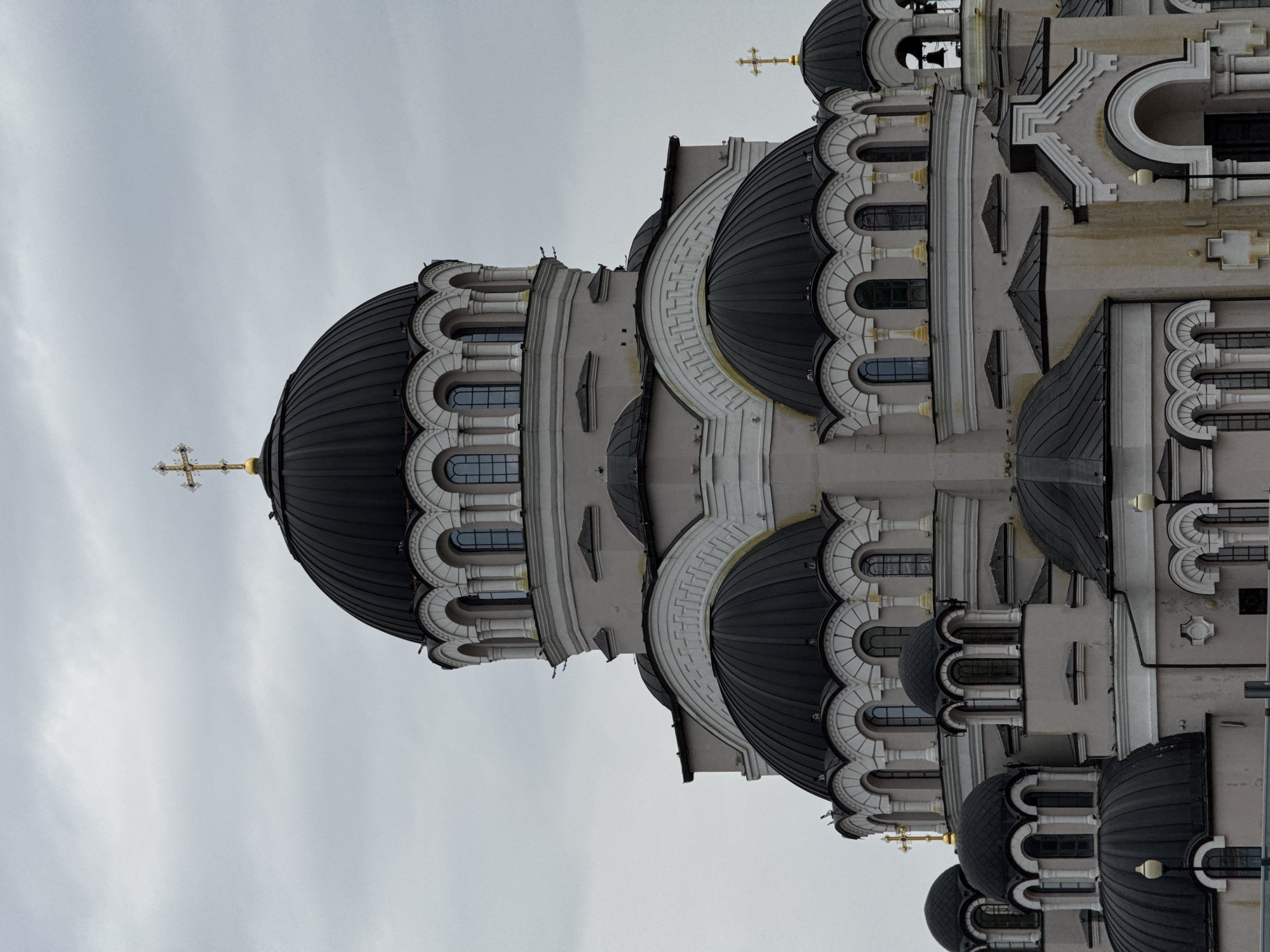 This image captures the exterior of a majestic Orthodox church or cathedral, seen under an overcast, grey sky. The grand religious edifice features numerous dark grey or black domes, several of which are topped with ornate golden Orthodox crosses. A prominent large dome with a golden cross dominates the left side of the frame (when oriented correctly), while smaller domes and architectural elements extend across the rest of the building.
The church's walls are a light beige or grey, richly adorned with intricate detailing. These details include multiple tiers of arched windows, many encased in elaborate scalloped or radially patterned surrounds. Various architectural embellishments like columns, pilasters, and patterned friezes (one large arch displays a distinct geometric pattern) contribute to the ornate and layered design. Bell towers are visible, with bells peeking from one section beneath a smaller dome.
The overcast sky suggests daytime with diffused lighting, casting no harsh shadows and highlighting the texture and form of the building's intricate facade. No people or specific activities are visible, placing full emphasis on the architectural grandeur of the structure. The style, particularly the onion domes and crosses, is characteristic of Eastern Orthodox churches found in regions like Sirius, Russia, as per the provided context. There is no visible text in the image.