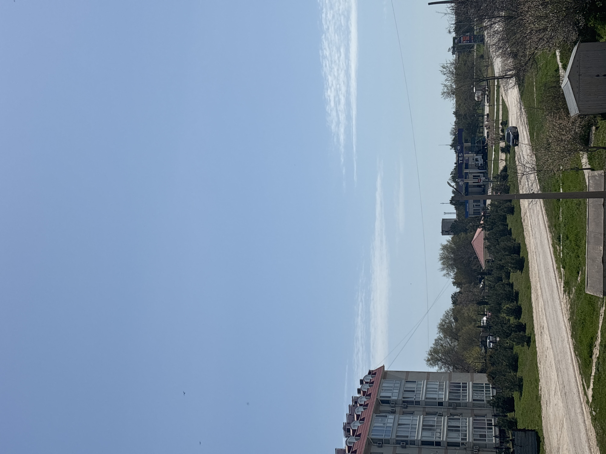 This outdoor image presents a daytime view of an area in Chornomorskoe, Ukraine, likely captured from an elevated position. The sky dominates the upper portion of the frame, a clear light blue with wispy white clouds stretching horizontally across the middle. Several small, dark specks, possibly birds, are visible against the sky.
On the left side, a multi-story building with a reddish, gabled roof and numerous large, white-framed windows is prominent. Many air conditioning units are visible beneath the windows of this building.
A dirt or gravel road runs through the midground, curving slightly and flanked by green grass and a variety of trees, some with bare branches, suggesting early spring. Along this road, further into the scene, a gas station with a blue canopy and "TES" clearly visible on its signage can be seen, with several cars present. Other structures, including a building with a red roof partially hidden by trees and a smaller shed-like building on the far right, are also part of the landscape. Utility poles and power lines extend across the scene. The weather appears sunny and clear, with bright daylight illuminating the area. No specific human activity is distinctly visible.